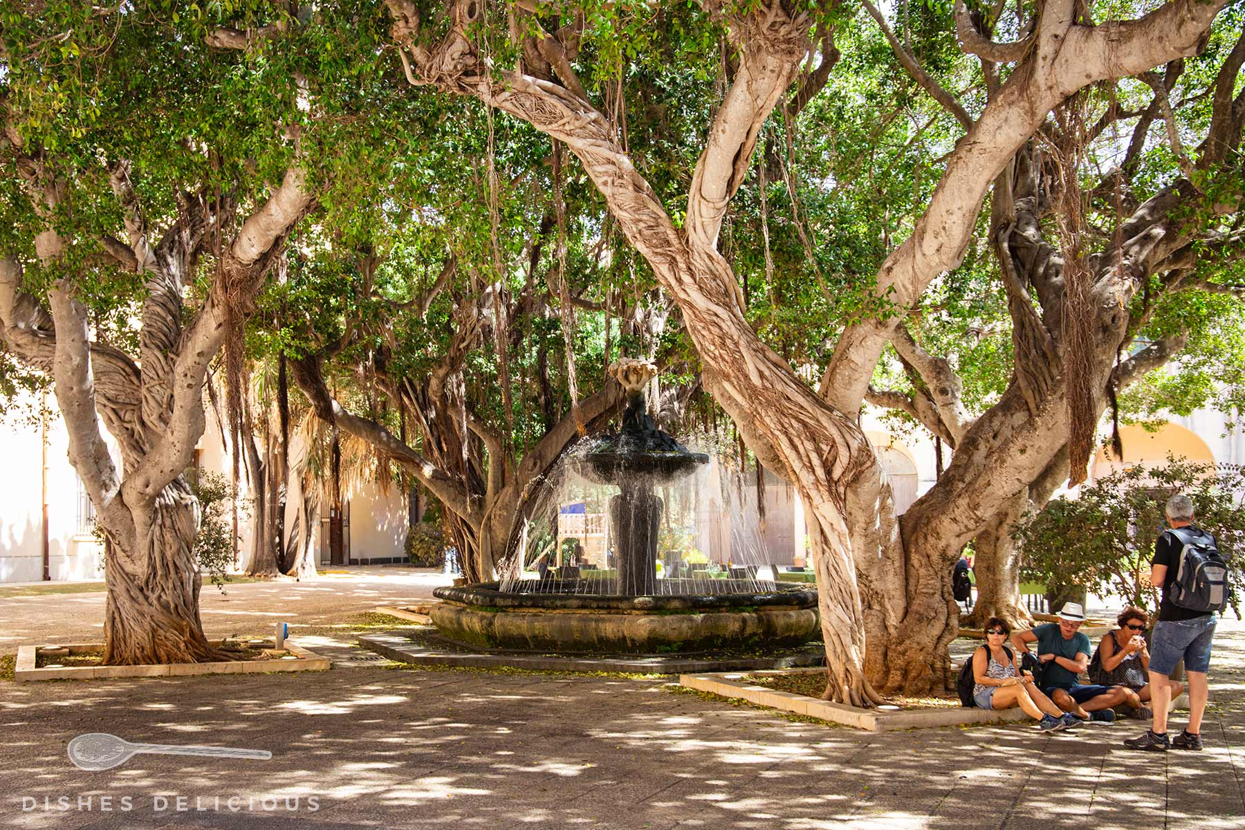 Plätschernder Brunnen in einem von großen Bäumen beschatteten Innenhof, daneben sitzt eine Gruppe von Menschen auf dem Boden.