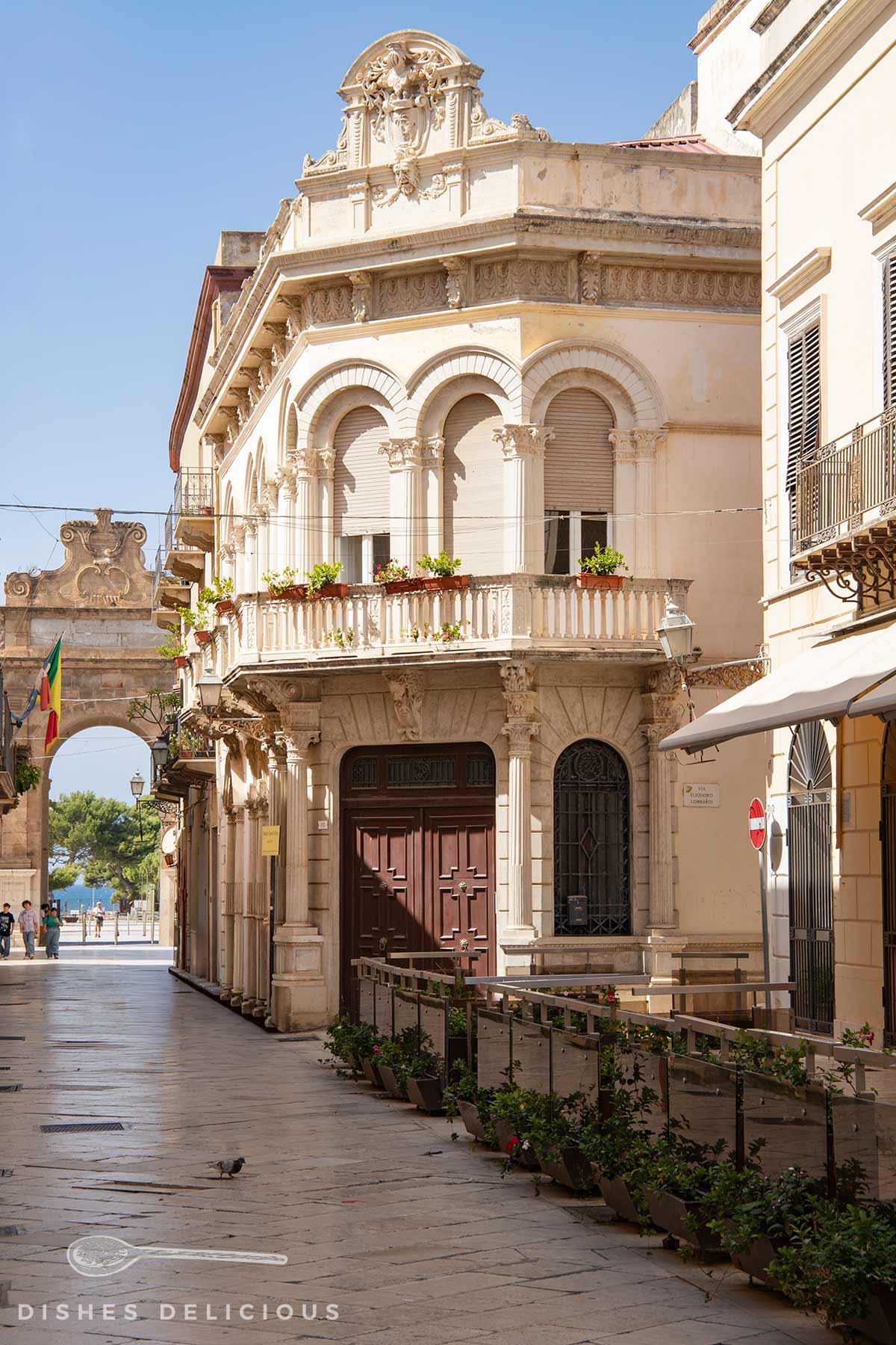 Palazzo Spano-Burgio mit verzierten Bögen und spanischem Balkon an einer sonnigen Straße, im Hintergrund ein Torbogen mit Flagge.