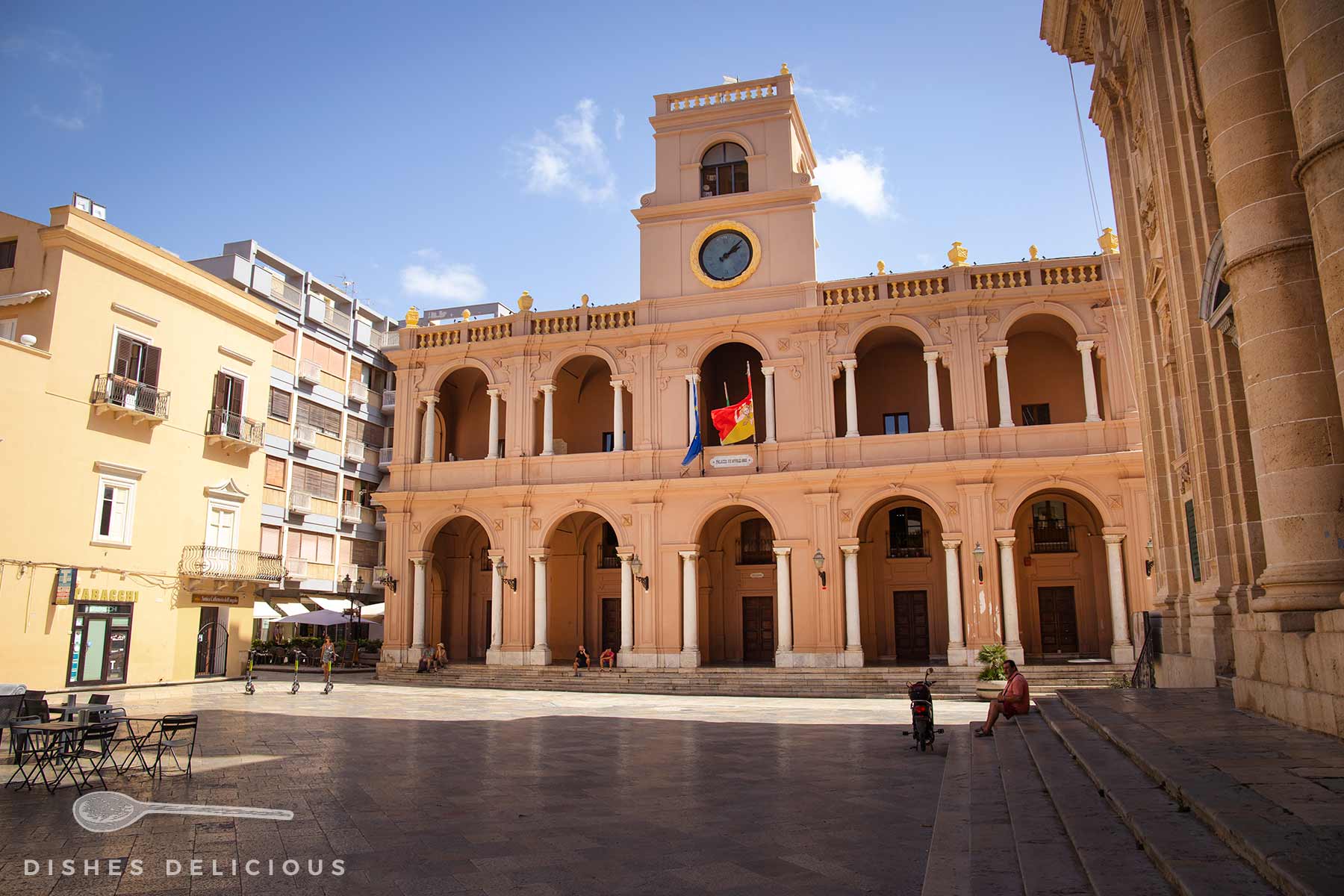 Fassade Palazzo VII Aprile in Marsala mit Uhrturm und Flagge auf einem sonnigen, fast menschenleeren Platz.