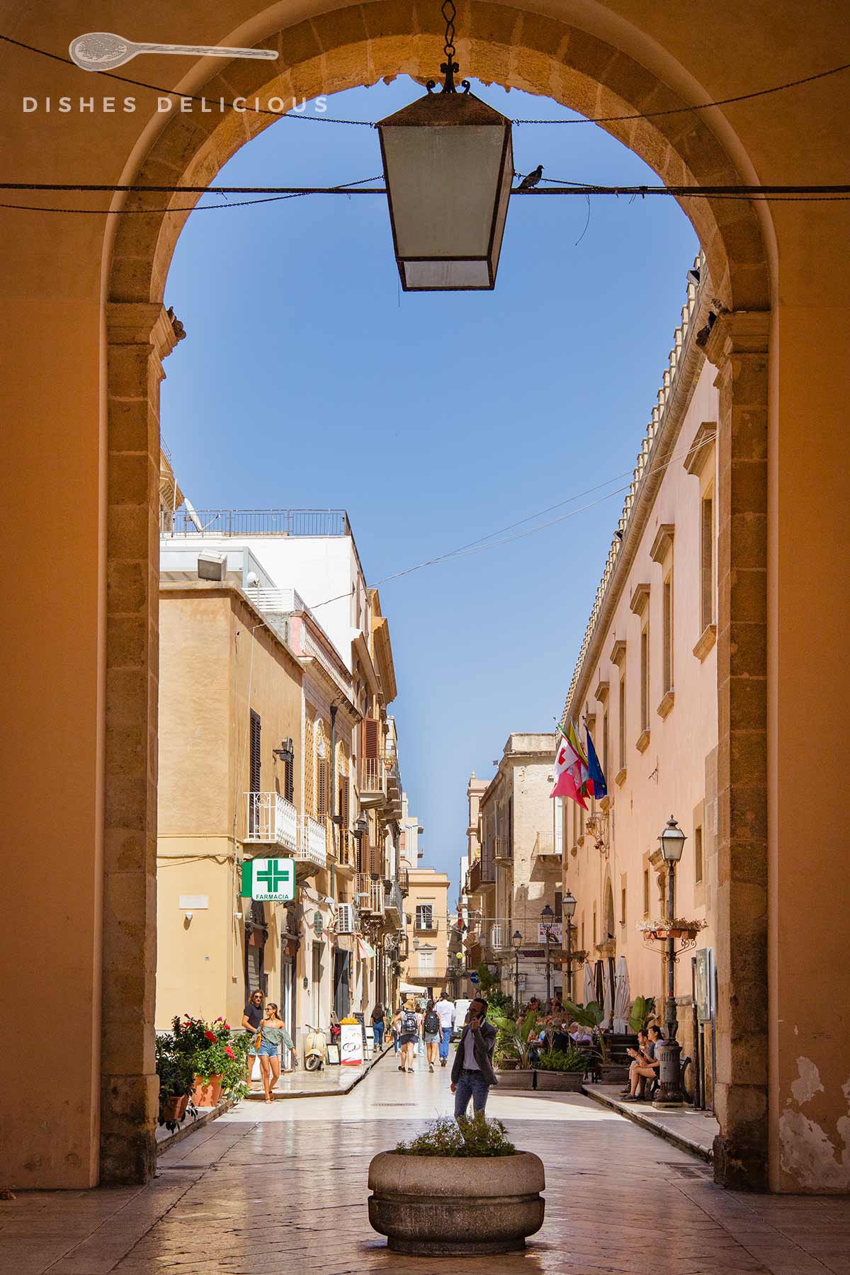 Blick durch einen steinernen Torbogen auf eine sonnige Fußgängerzone mit mehreren Gebäuden, Passanten und einer hängenden Laterne.