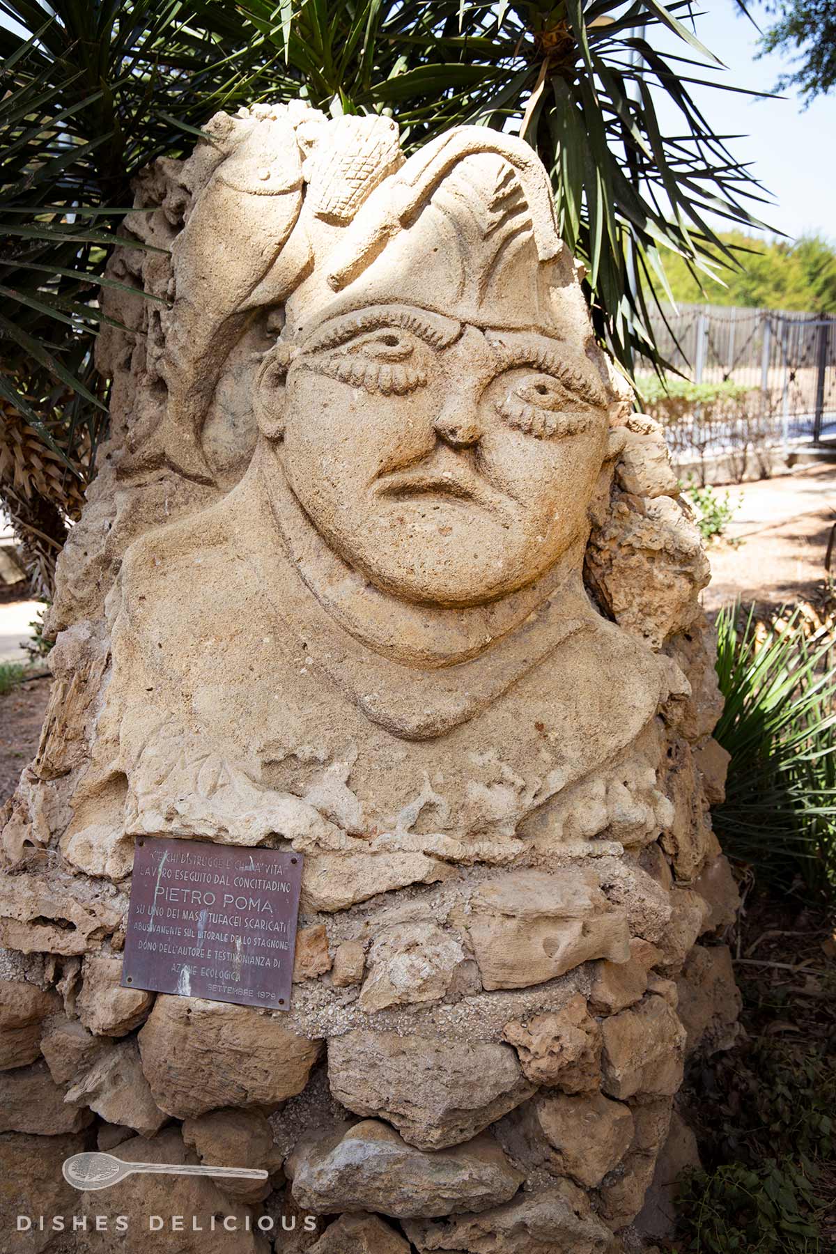 Sandsteinskulptur mit dekorativen Elementen und einer Metalltafel am Sockel, umgeben von Palmenblättern im Freien.
