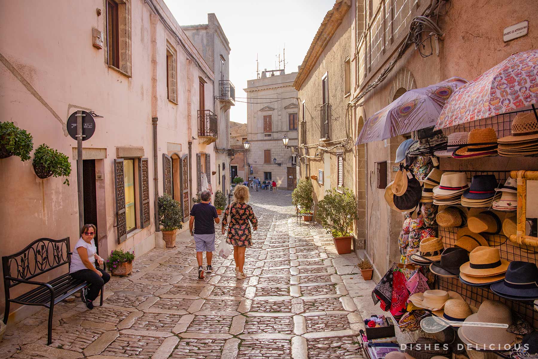 Schmale Gasse mit Kopfsteinpflaster, links Bank, auf der eine Frau sitzt, rechts Verkaufsstand mit Hüten und Regenschirmen, zwei Personen spazieren auf der Straße.