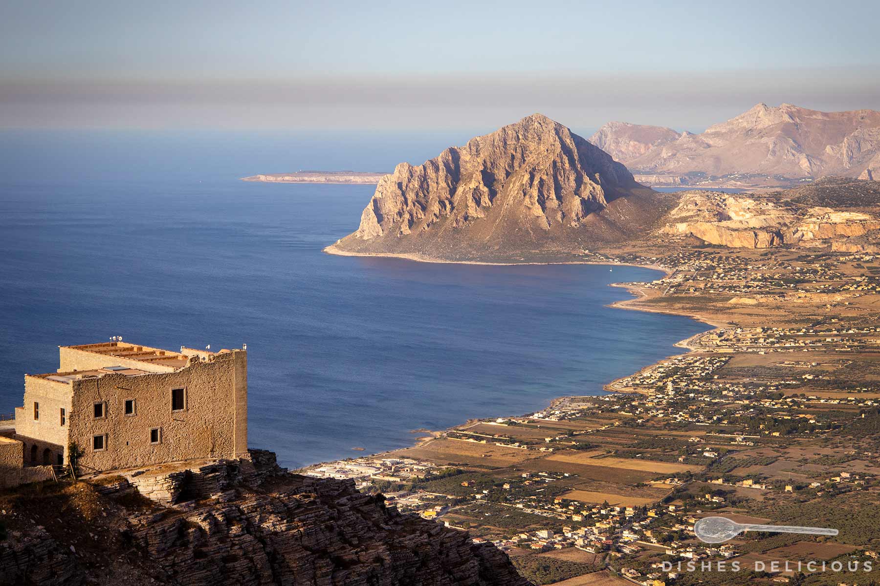 Blick auf die Küstenlandschaft unterhalb von Erice mit einem Gebäude auf einem Felsen im Vordergrund und einem großen Berg (Monte Cofano) am Meer im Hintergrund.