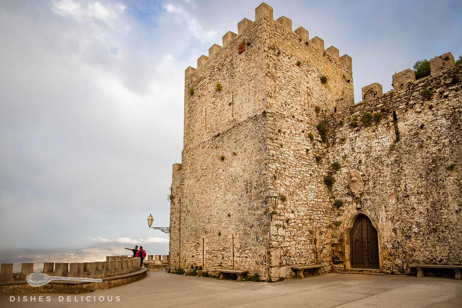Castello del Balio: Großer mittelalterlicher Steinturm mit Zinnen und Holztür, davor zwei Personen an einer Steinmauer unter bewölktem Himmel.