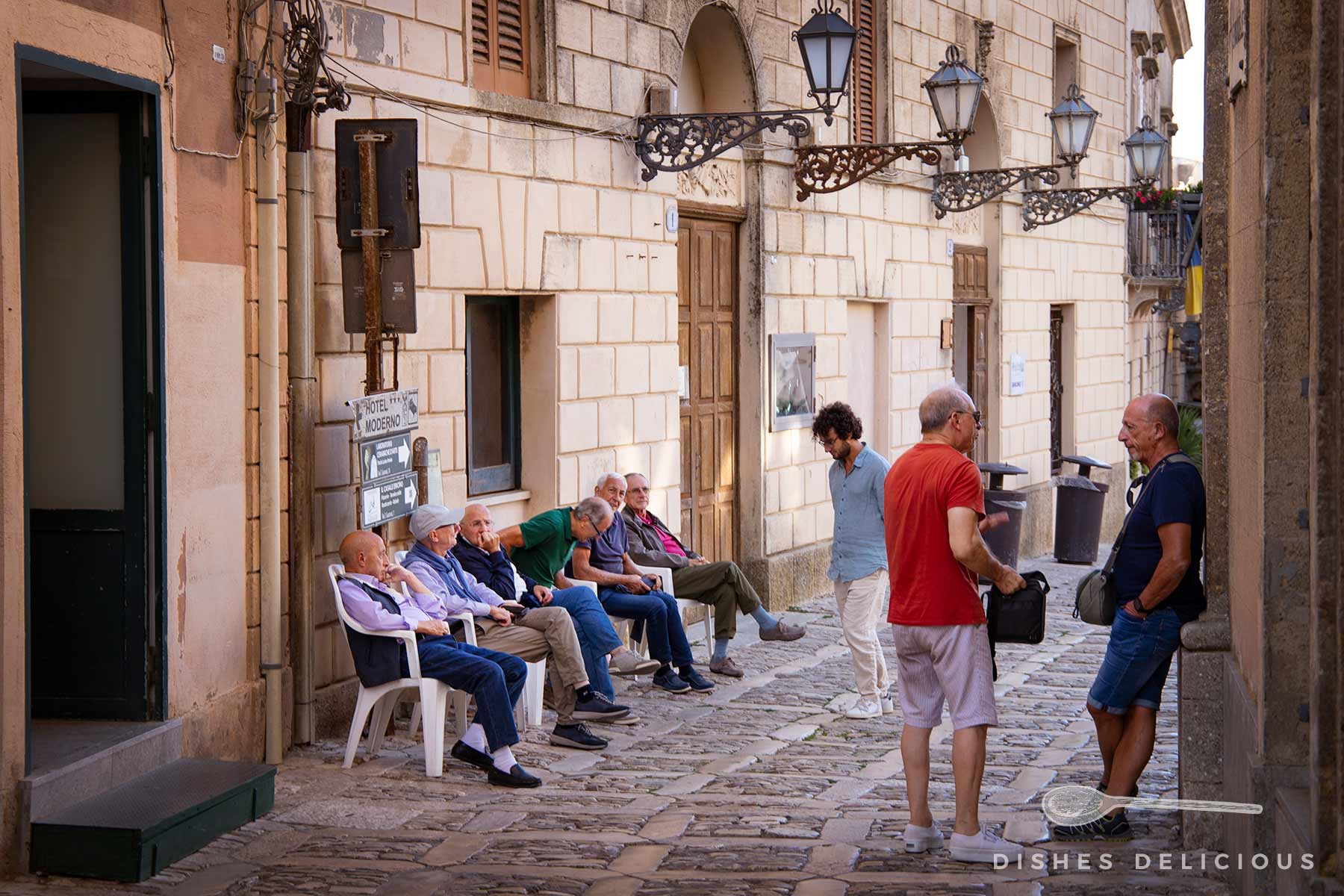 Mehrere ältere Menschen sitzen auf Stühlen und unterhalten sich in einer gepflasterten Gasse mit historischen Gebäuden und Laternen.