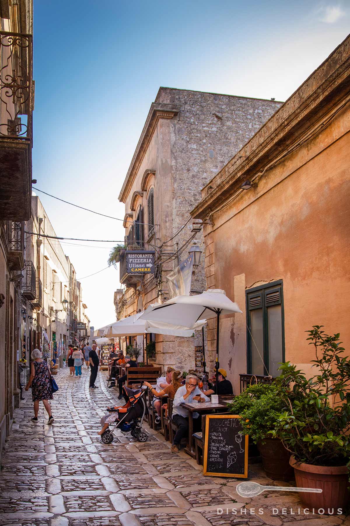 Belebte Kopfsteinpflastergasse mit Außentischen eines Restaurants, Sonnenschirmen und Passanten im Centro Storico von Erice unter blauem Himmel.