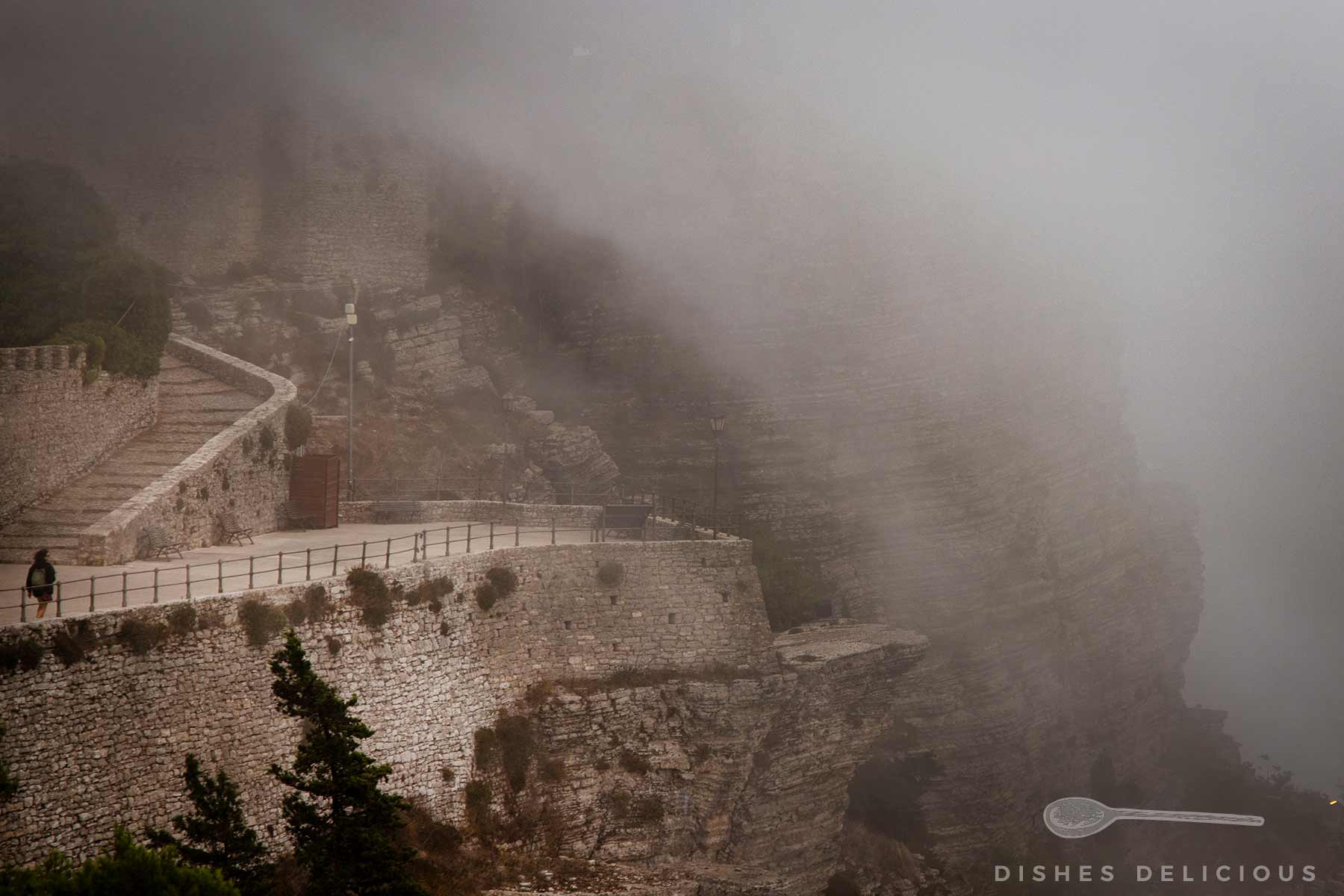Nebelschwaden umhüllen eine steinerne Mauer und einen Weg an einem Felsen, eine einzelne Person geht am Geländer entlang.