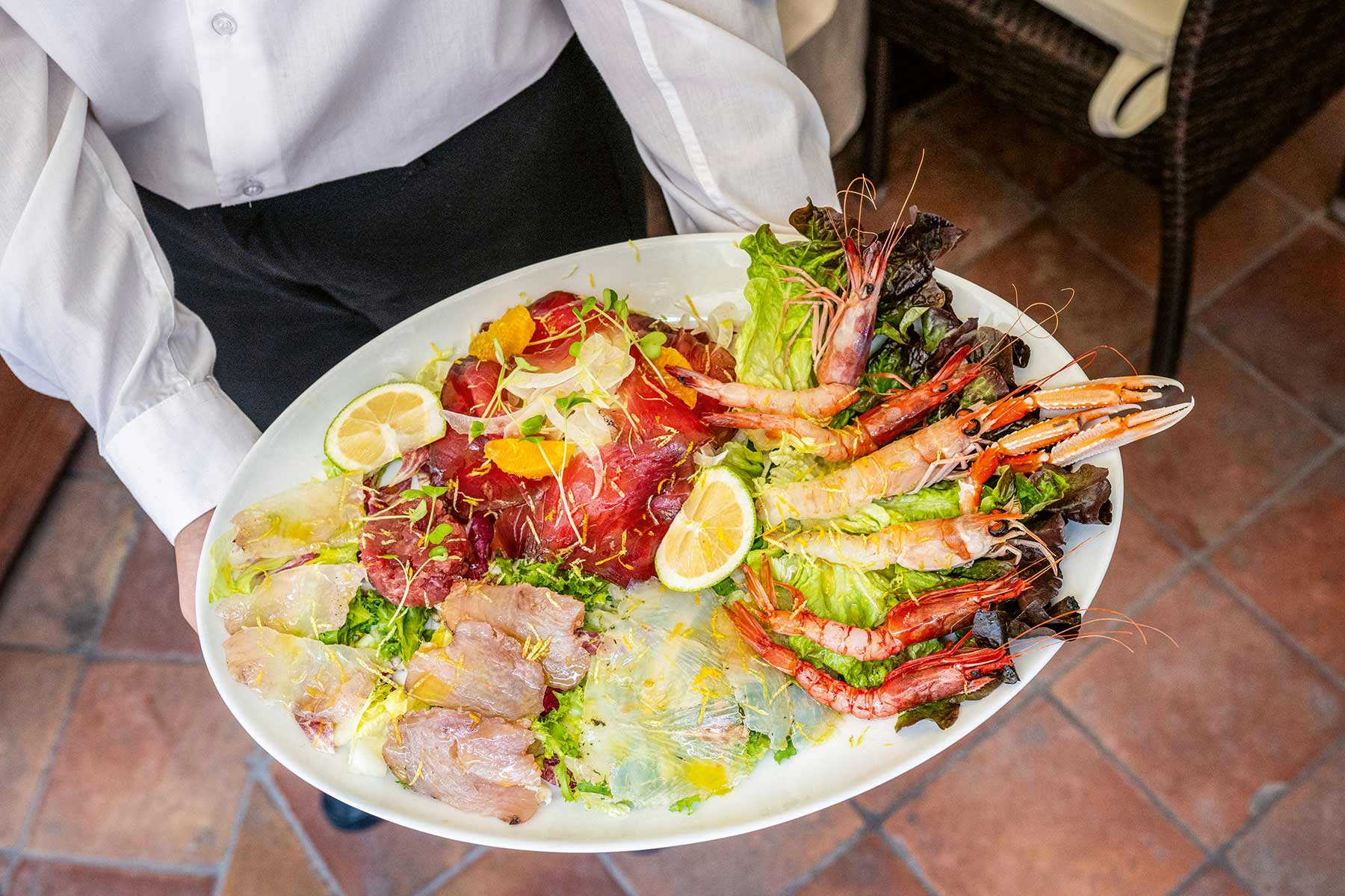 Servierperson trägt einen großen weißen Teller mit rohem Fisch, Garnelen, Salat und Zitronenscheiben.