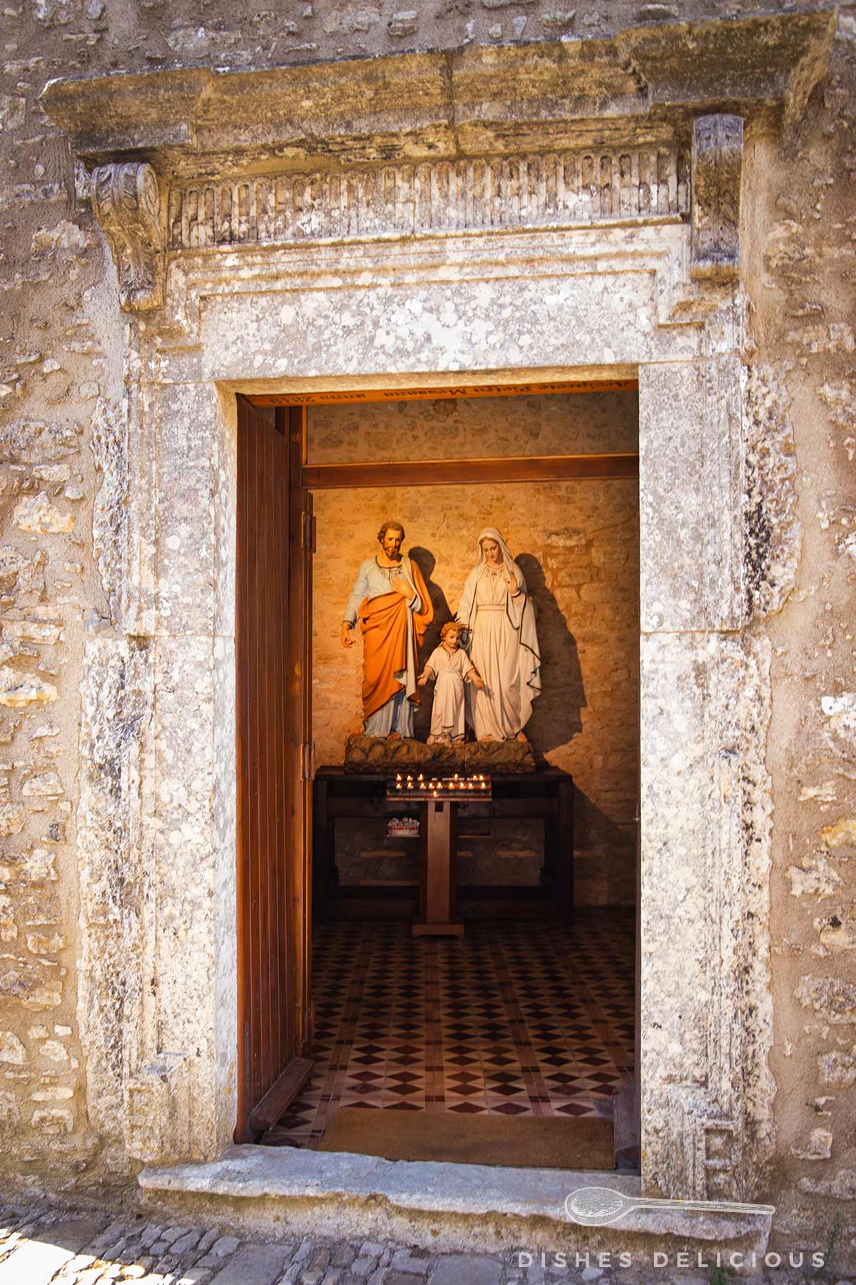 Offene Tür mit steinerner Umrahmung führt zu einer Kapelle mit einer religiösen Statue von drei Figuren auf einem Podest und brennenden Kerzen davor.