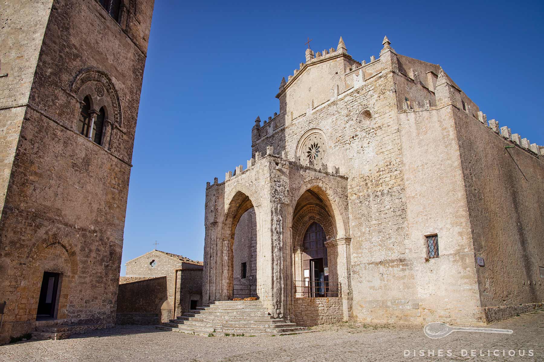 Kathedrale von Erice, erbaut aus hellem Stein mit romanischen Bögen und Rosettenfenster bei blauem Himmel. Links der Glockenturm.