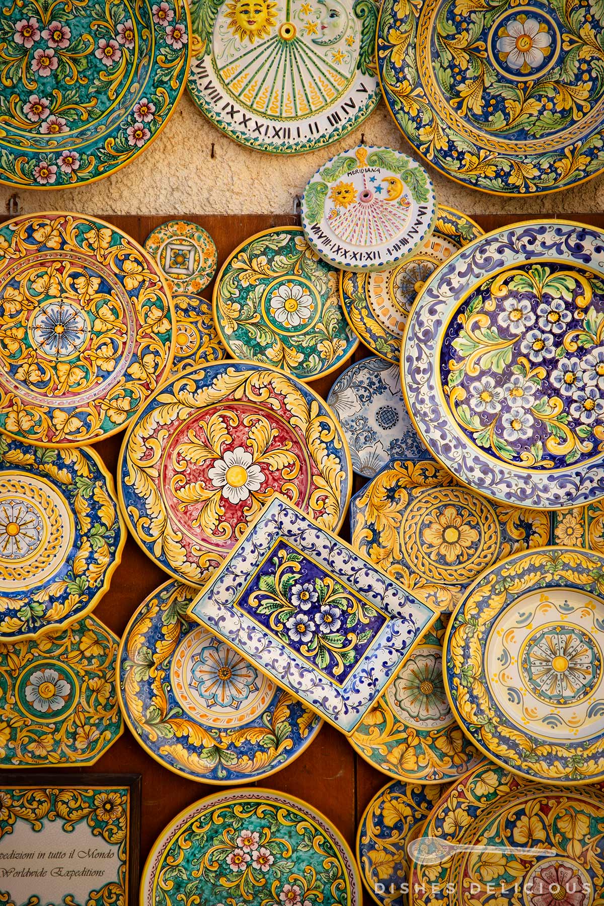 Bunte, handbemalte Keramikteller mit floralen und ornamentalen Mustern sizilianischer Art, darunter ein rechteckiger Teller mit blau-lila Blumenmotiven.