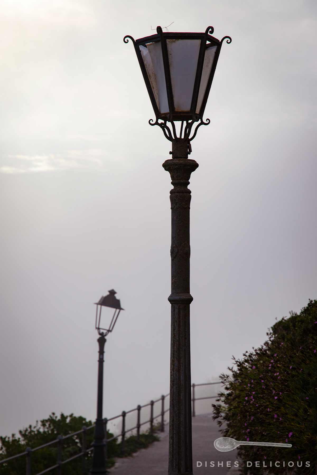 Alte Straßenlaterne aus Metall mit verzierten Elementen vor nebligem Himmel und einer weiteren Laterne im Hintergrund.
