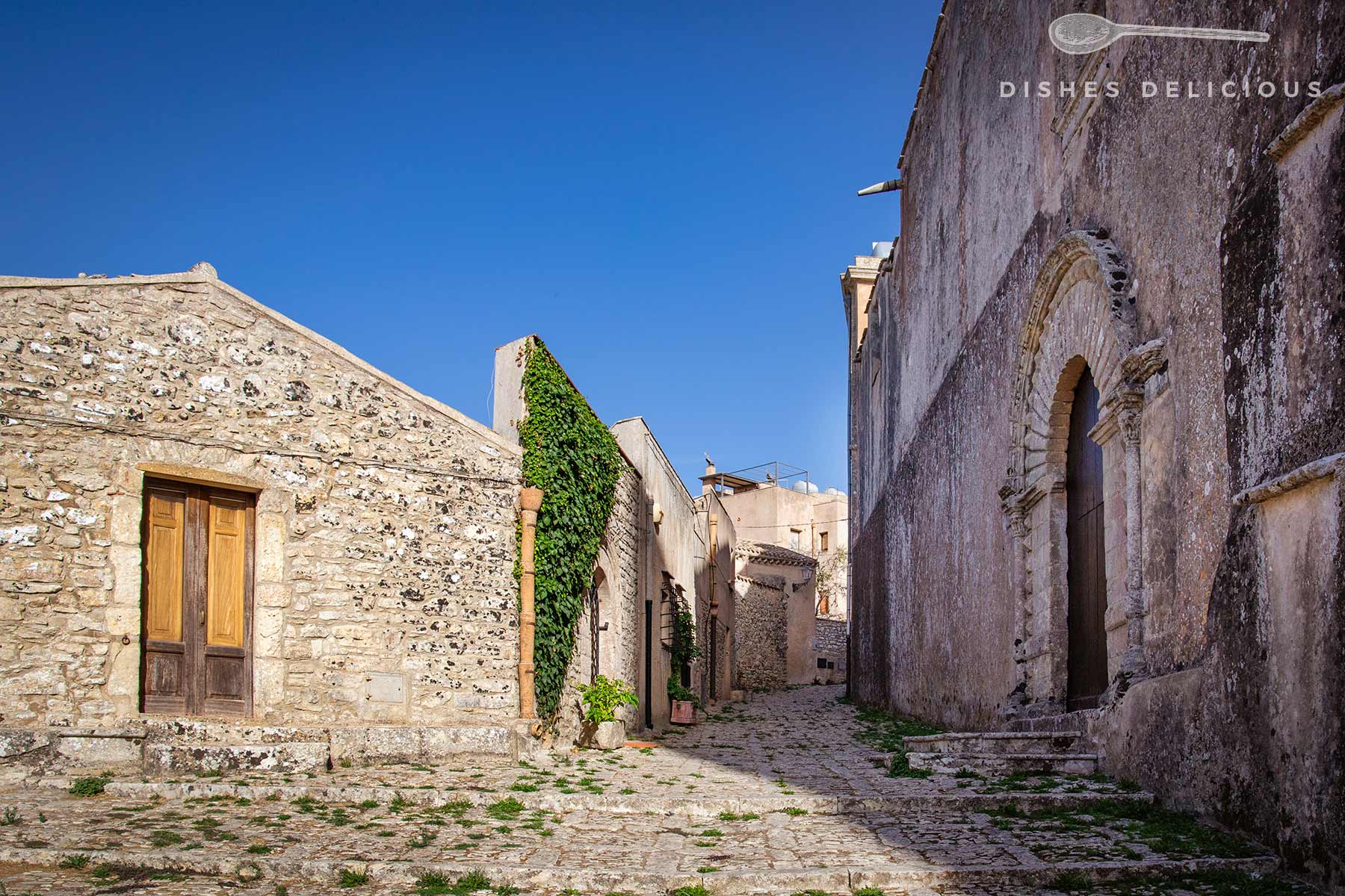 Schmaler gepflasterter Weg zwischen alten Steinbauten mit Holztüren und Efeu an der Wand unter blauem Himmel.