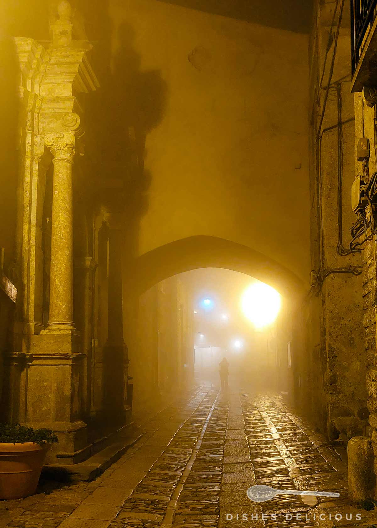 Nebelige Gasse in Erice mit Kopfsteinpflaster am späten Abend und einer Person im Lichtschein am Ende des Wegs.