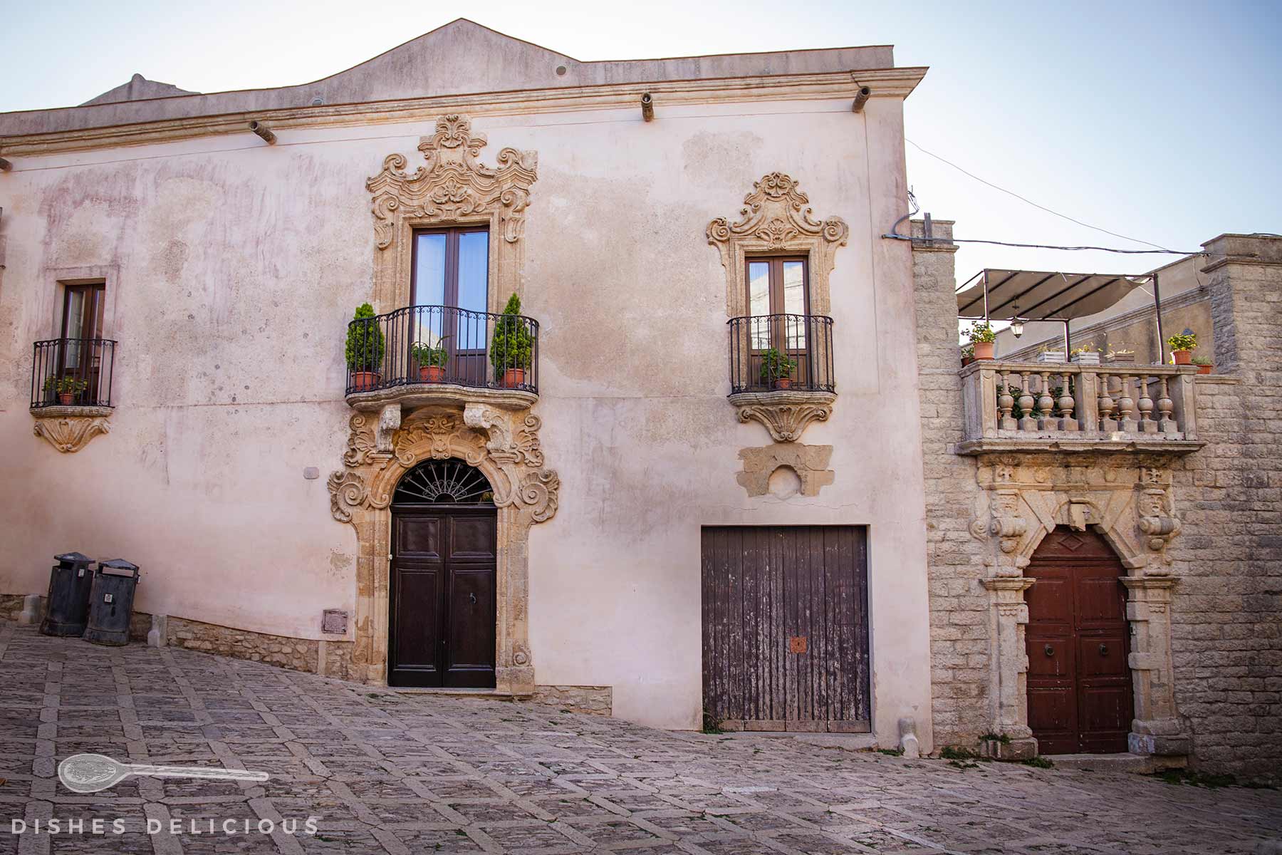 Fassade eines historischen Hauses mit verzierten Fenstern, einem Balkon und zwei Türen an einer gepflasterten Straße.