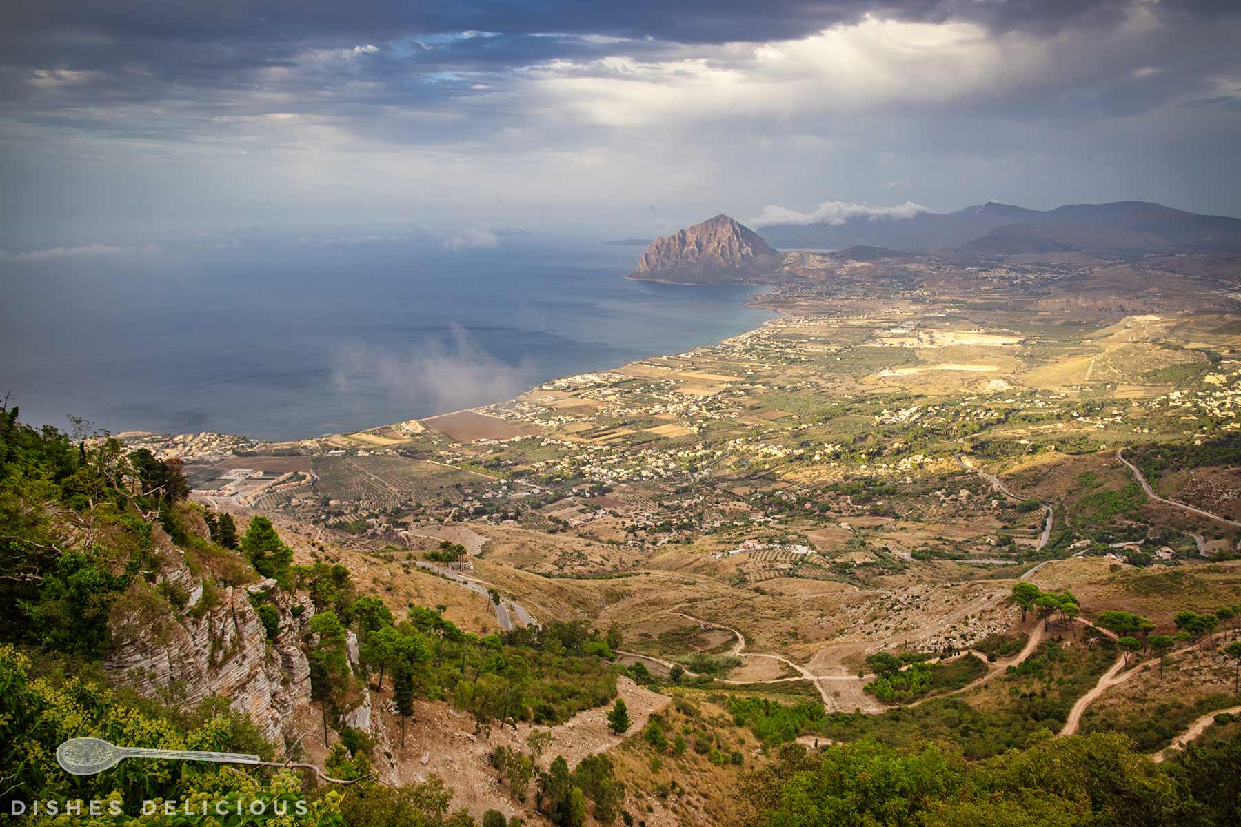 Blick vom Monte Erice auf die Küstenlandschaft mit Feldern, Wegen und einem markanten Berg am Meer.