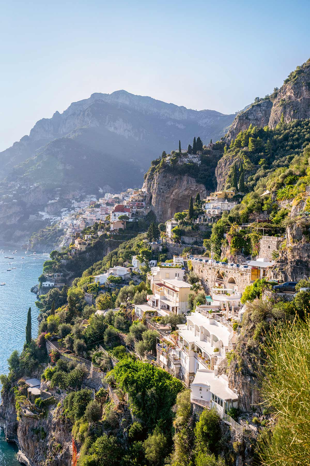 Küstenort mit terrassenförmig angeordneten Häusern an steilen Klippen über dem Meer und bewaldeten Bergen im Hintergrund.