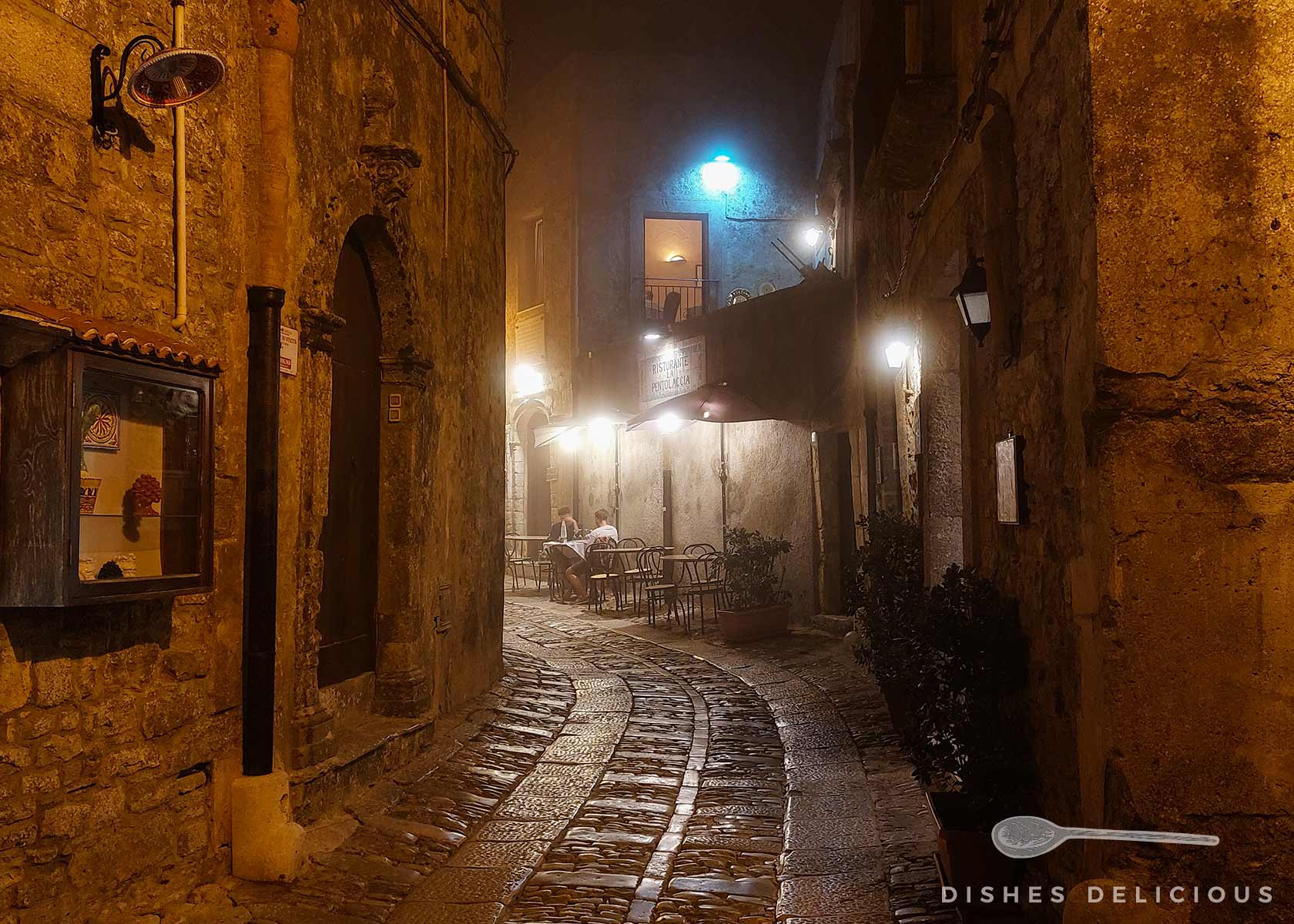 Beleuchtete Kopfsteinpflastergasse mit Tischen und Stühlen vor einem Restaurant in einem alten Steingebäude bei leicht nebeliger Nacht.