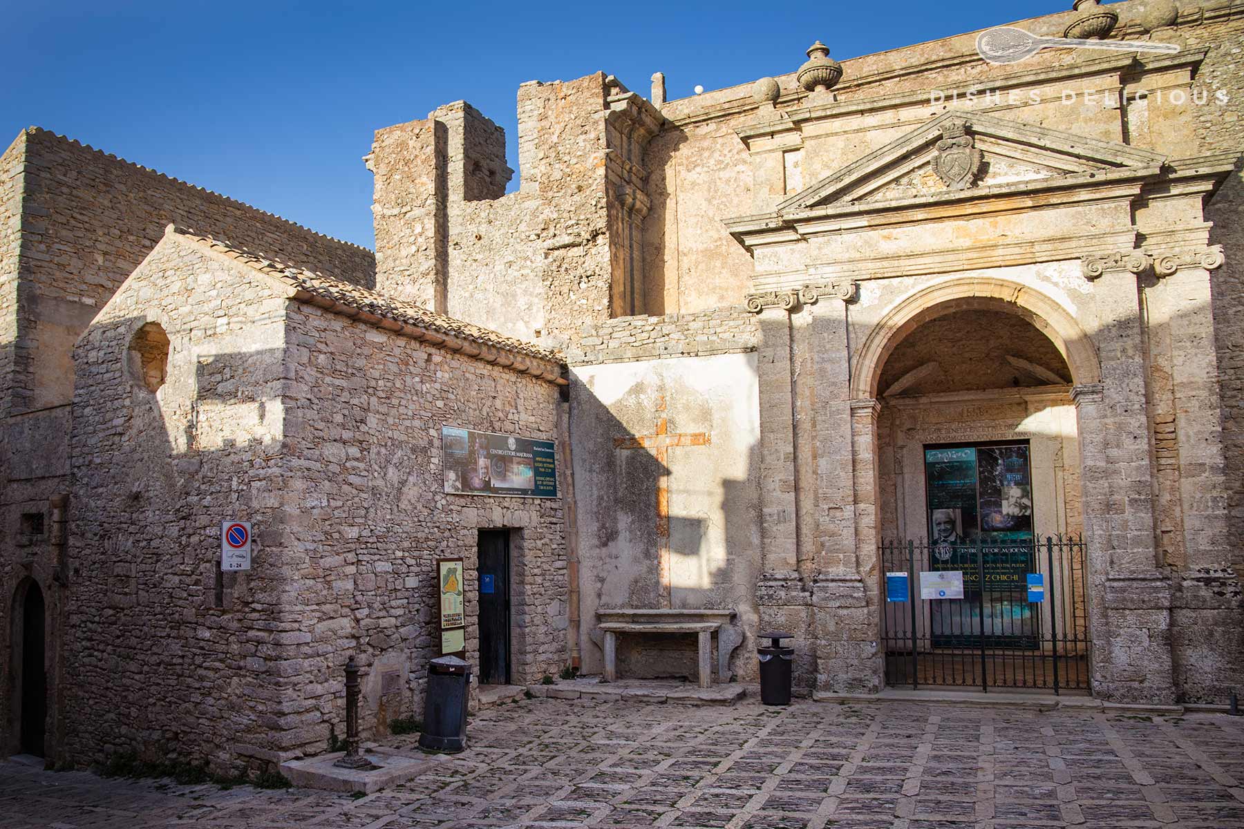 Steinerne Fassade eines historischen Gebäudes mit Rundbogenportal und kleiner Kirche, Pflaster aus Naturstein.