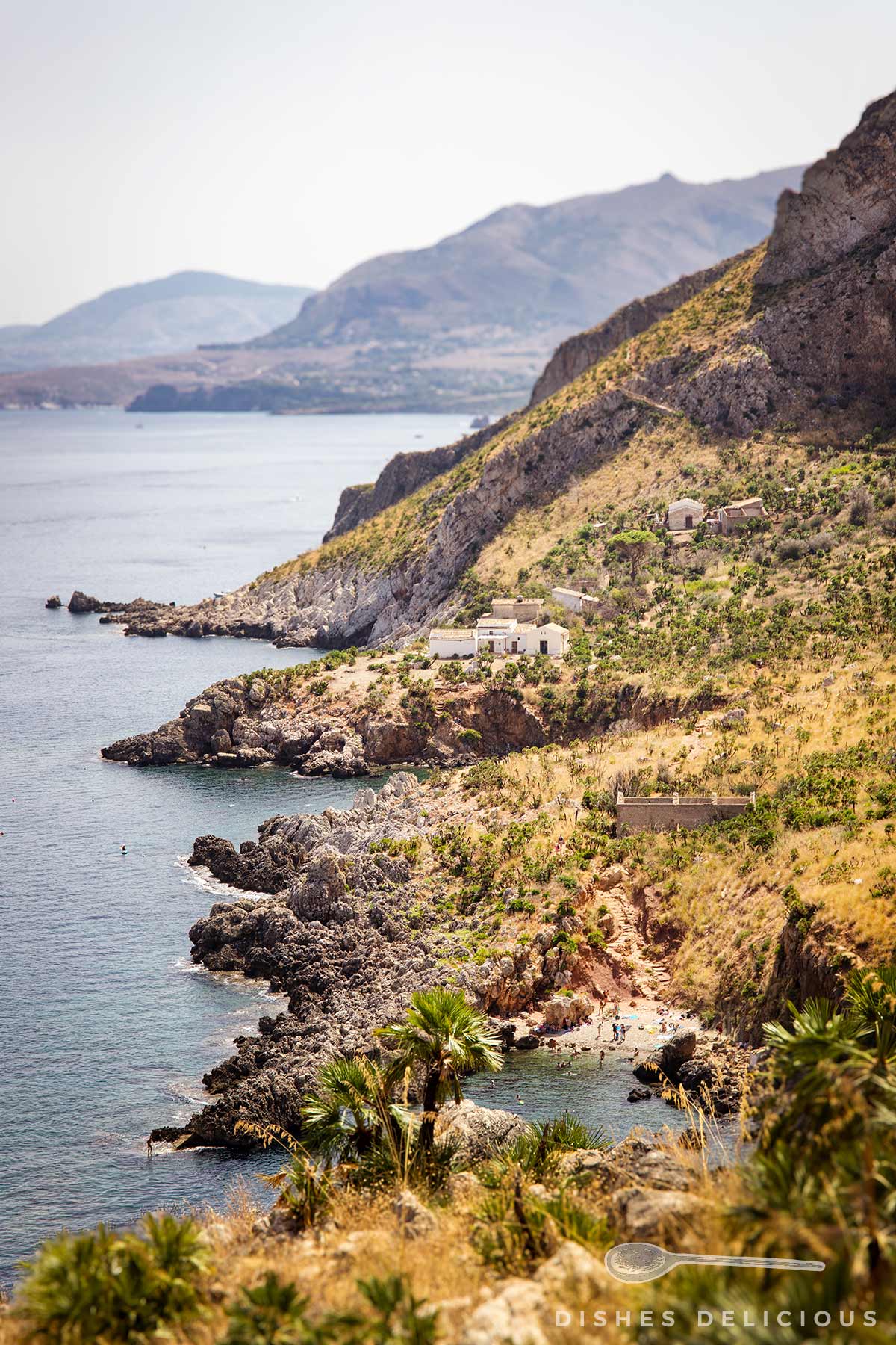 Küstenlandschaft des Lo Zingaro mit felsigen Klippen, vereinzelten Gebäuden und Vegetation vor einem ruhigen Meer und Bergen im Hintergrund