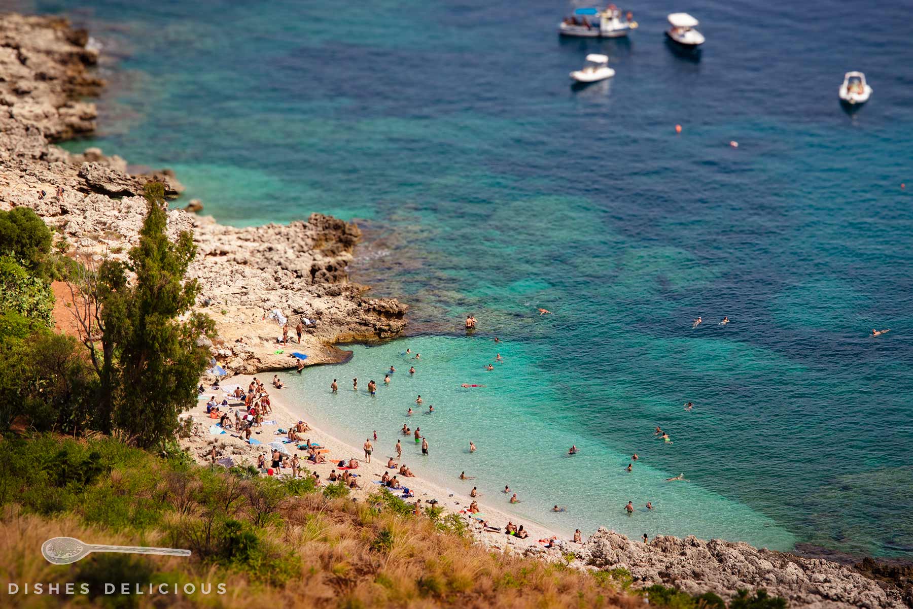 Kleine Bucht mit Sandstrand, umgeben von Felsen und klarem blauem Wasser, mit mehreren Menschen im Wasser und am Strand sowie einigen Booten im Hintergrund.