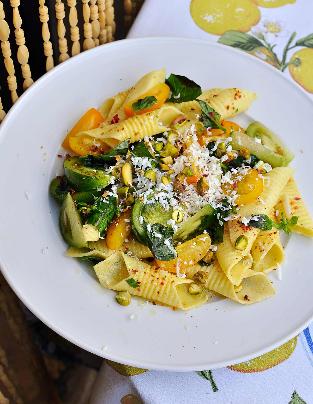 Ein Teller mit Garganelli-Nudeln, gelben und grünen Tomaten, Pistazienstückchen, Minzblättern und geriebenem Käse auf einem Tisch.