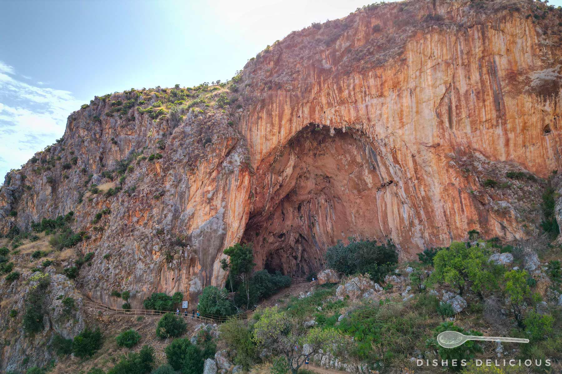 Große Felswand mit einer weit geöffneten Höhle und Vegetation am Fuß des Felsens.
