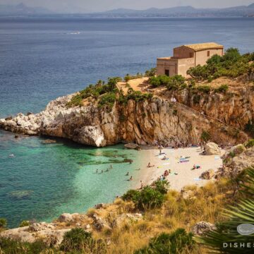 Kleine Bucht mit Sandstrand, umgeben von Felsen und Vegetation, mit einem Haus auf der Klippe und Menschen am Strand und im Wasser.