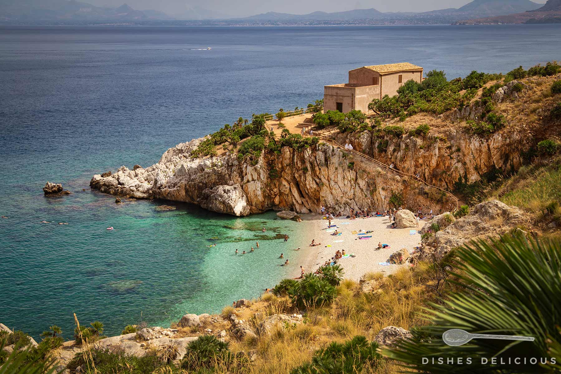 Kleine Bucht mit Sandstrand, umgeben von Felsen und Vegetation, mit einem Haus auf der Klippe und Menschen am Strand und im Wasser.