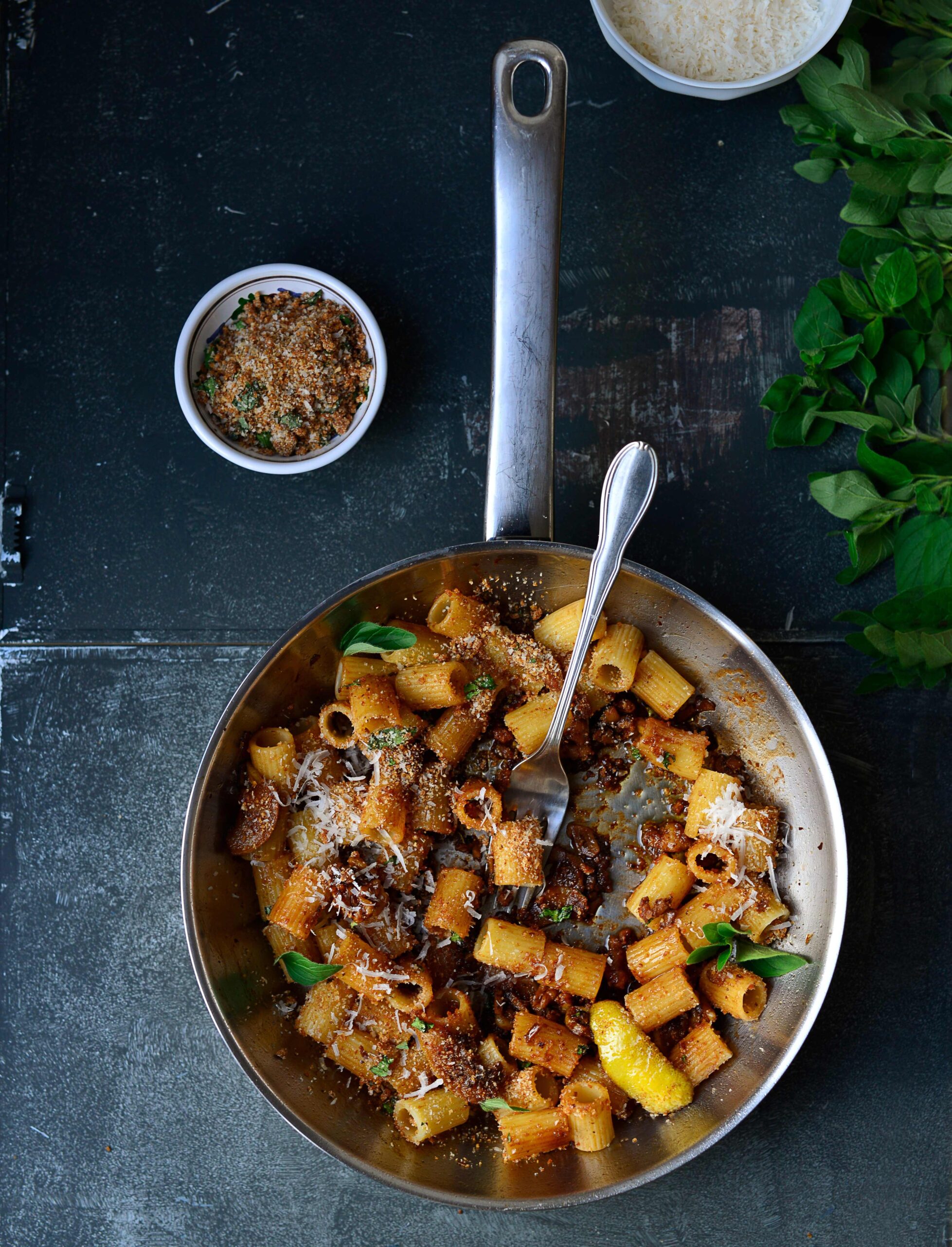 Pfanne mit Rigatoni in Pilz-Sardellen-Birnensauce auf dunklem Untergrund, daneben kleine Schale mit geriebenem Käse und Schale mit Umami-Bröseln.