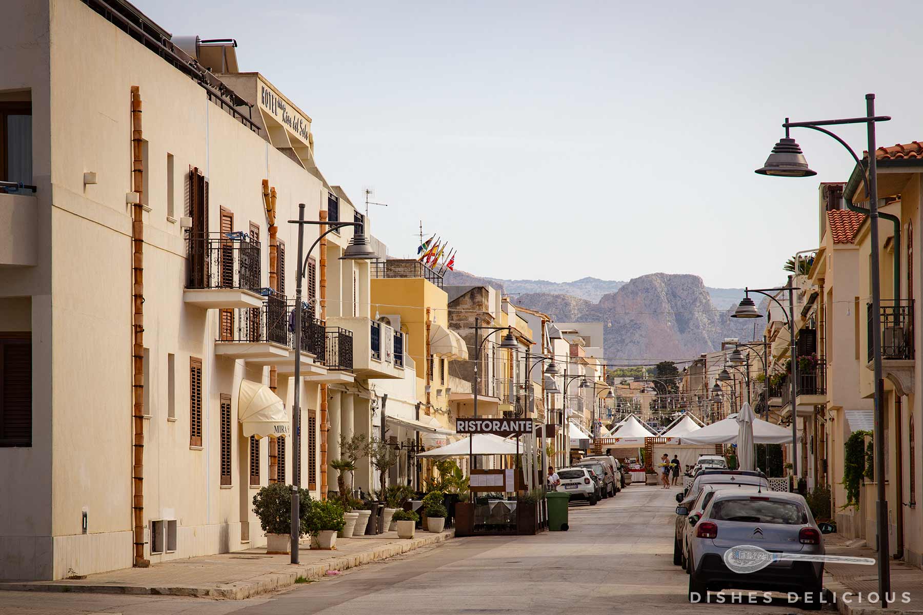 Straße in San Vito Lo Capo mit niedrigen Gebäuden, geparkten Autos und Bergen im Hintergrund.