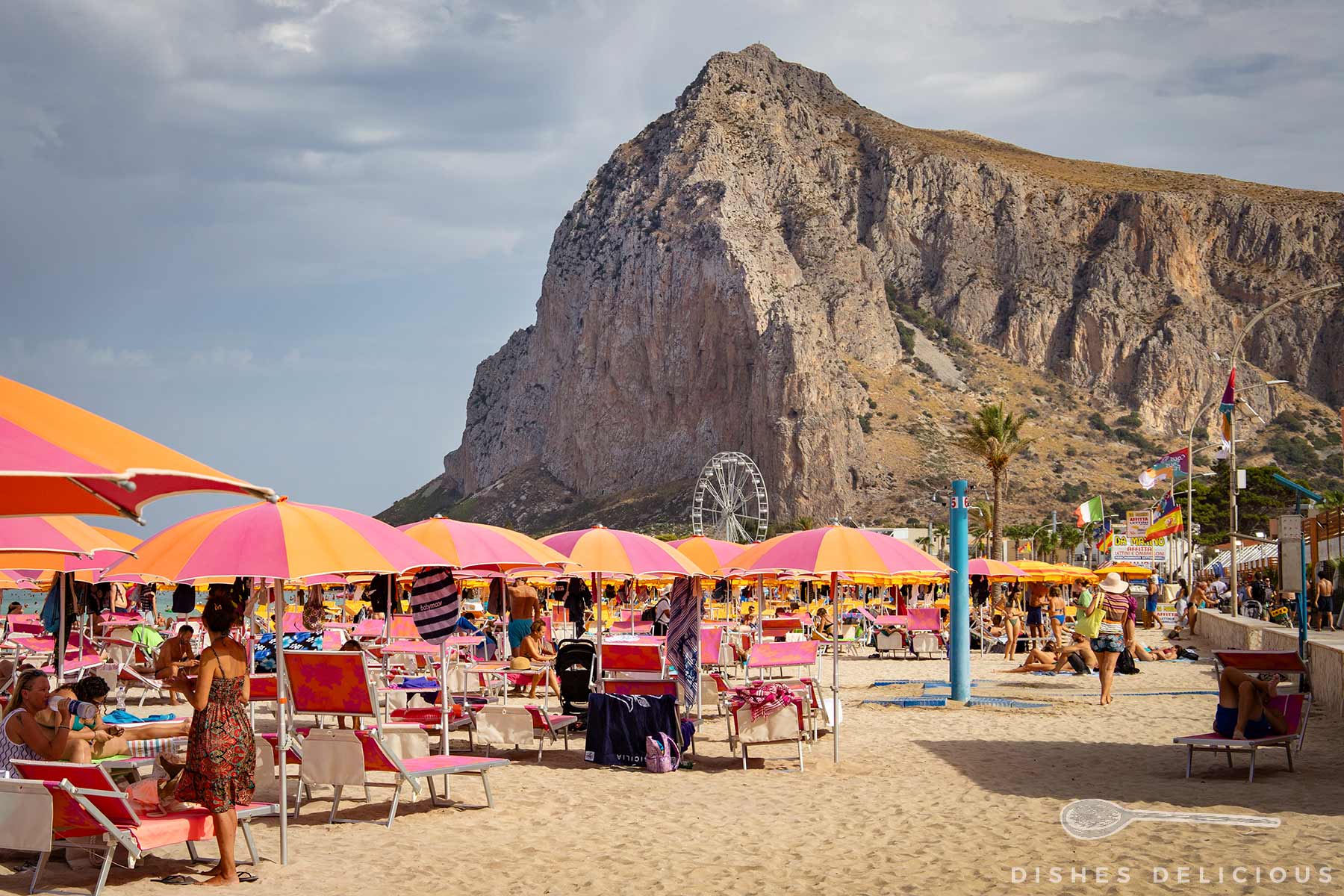 Strand mit vielen bunten Sonnenschirmen und Liegestühlen vor einem großen, felsigen Berg und einem Riesenrad im Hintergrund.