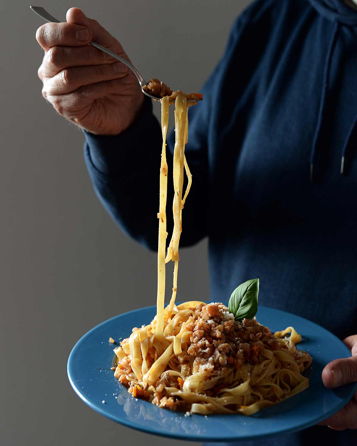 Jemand hält eine Gabel mit langen Tagliatelle über einem blauen Teller mit Pasta und einer Basilikumblättern als Garnitur.