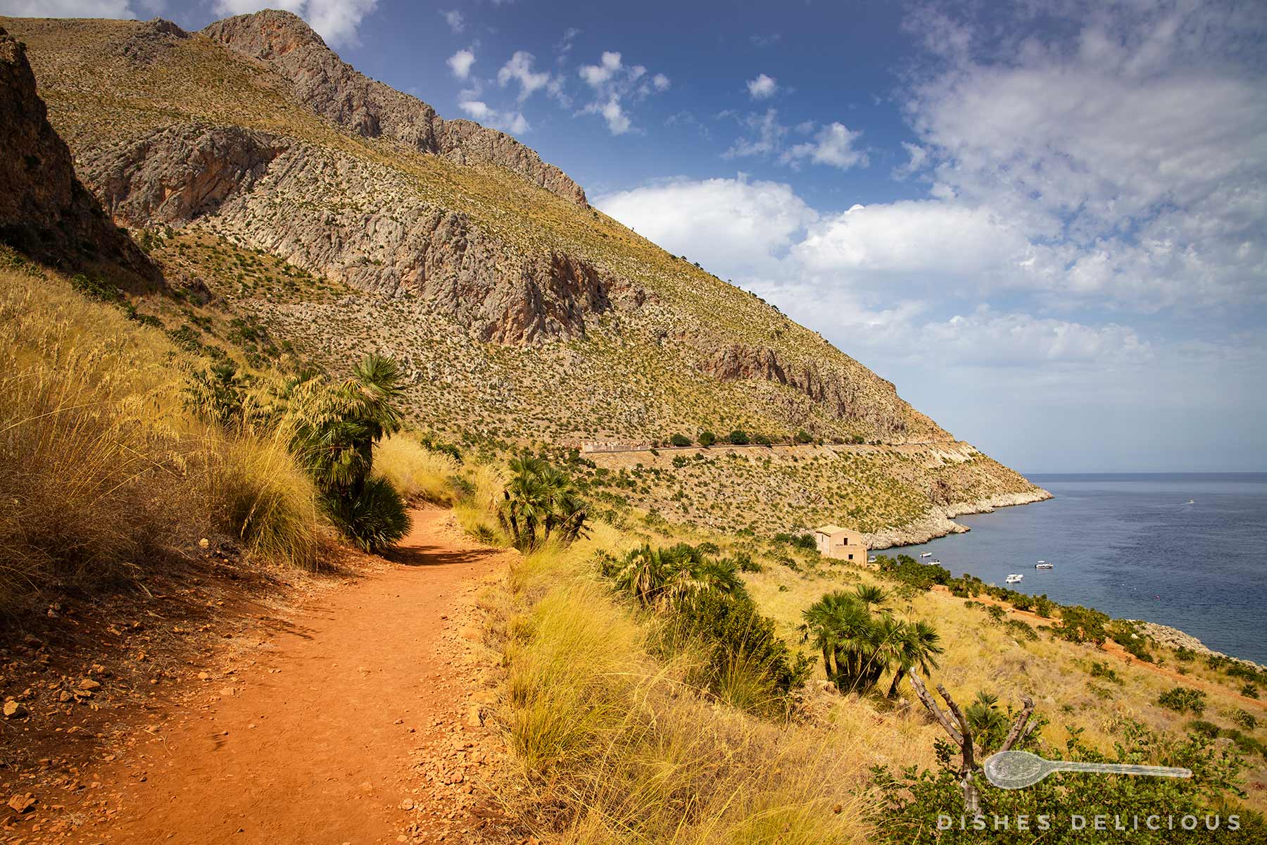 Wanderweg mit rotem Boden führt an trockenem Gras und Palmen vorbei zu einem kargen, felsigen Küstenhang mit Blick auf das Meer und Boote.