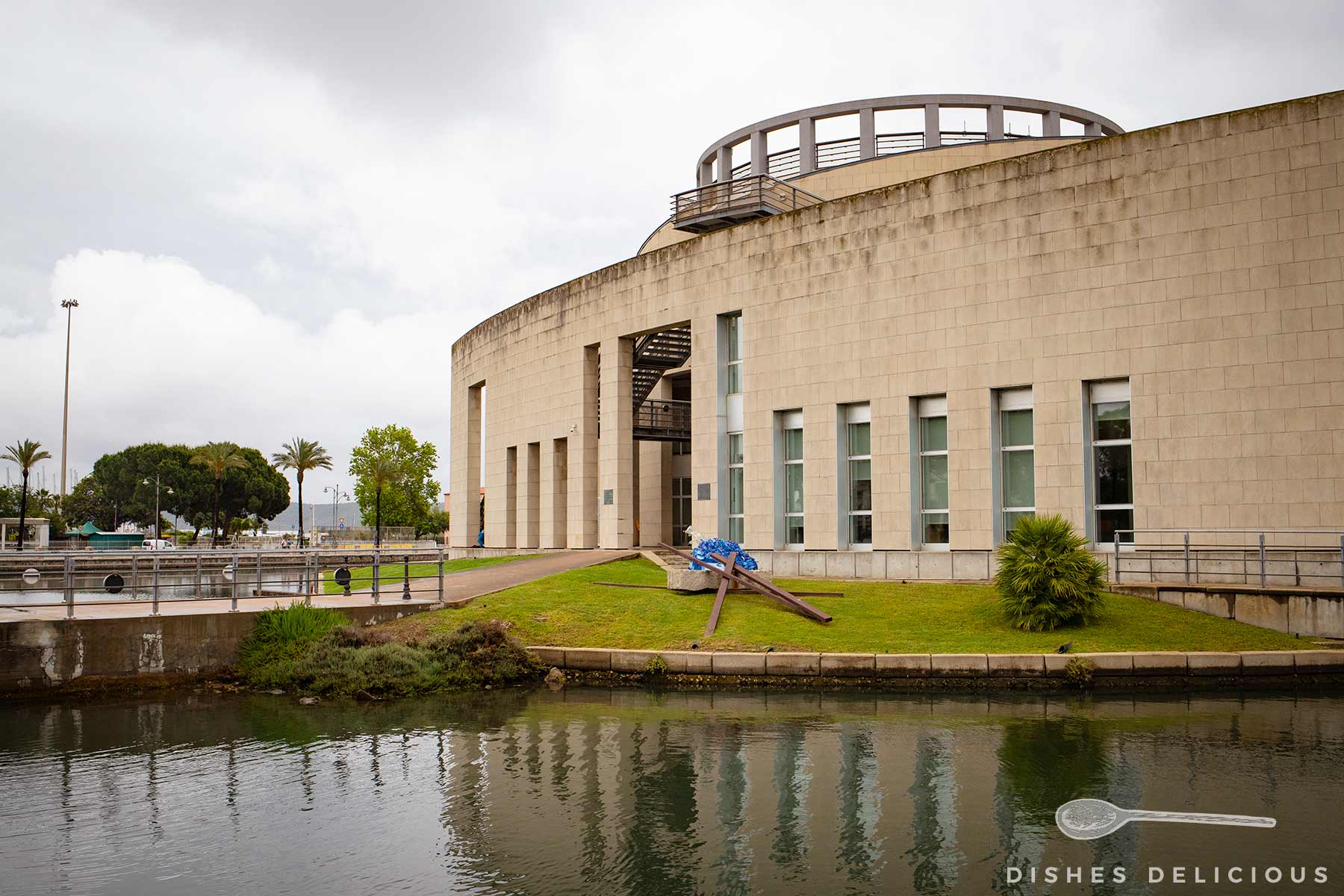 Rundes, modernes Museum mit Säulen und großen Fenstern an einem Wassergraben unter bewölktem Himmel.