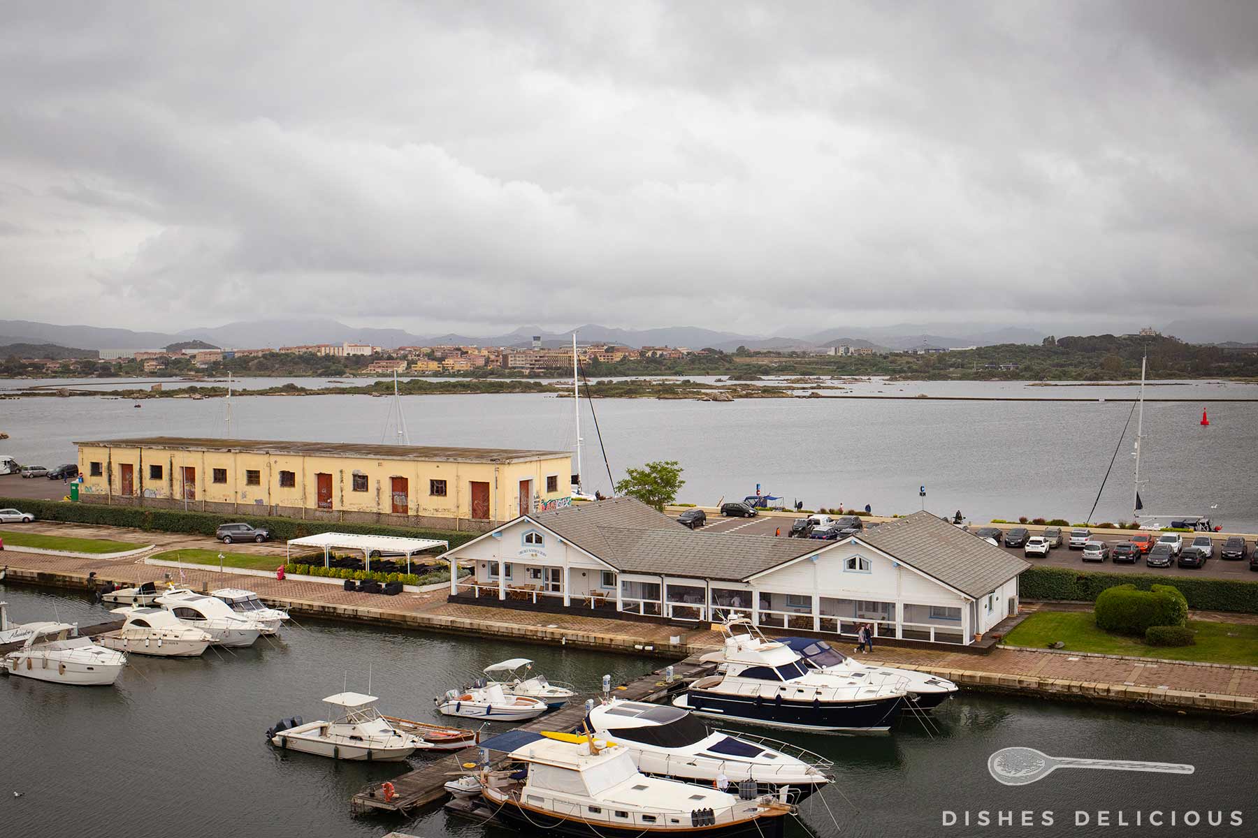 Blick auf einen kleinen Hafen mit mehreren Yachten am Golf von Olbia, einem weißen Gebäude am Wasser unter bewölktem Himmel.