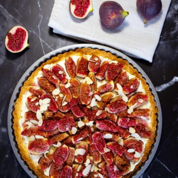 Crostata mit Feigen und Mandelblättchen belegt auf dunklem Untergrund neben zwei ganzen und zwei halbierten Feigen auf weißem Tuch.