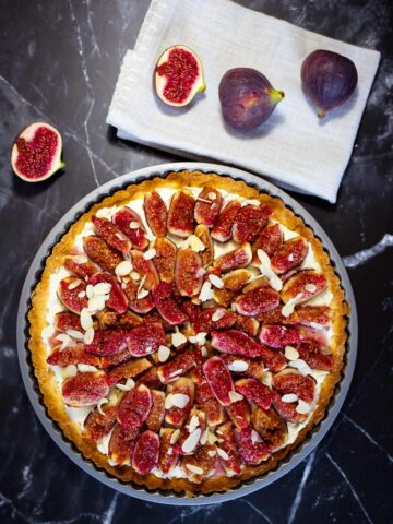 Crostata mit Feigen und Mandelblättchen belegt auf dunklem Untergrund neben zwei ganzen und zwei halbierten Feigen auf weißem Tuch.