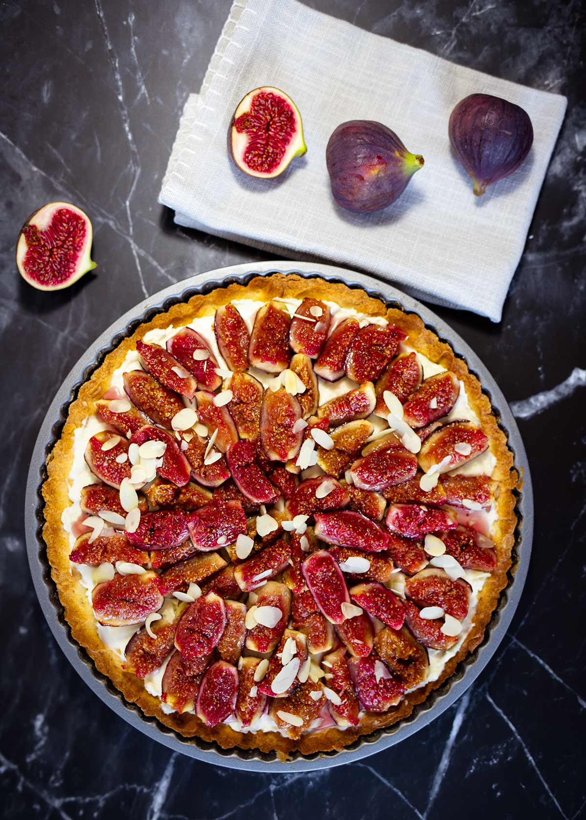 Crostata mit Feigen und Mandelblättchen belegt auf dunklem Untergrund neben zwei ganzen und zwei halbierten Feigen auf weißem Tuch.