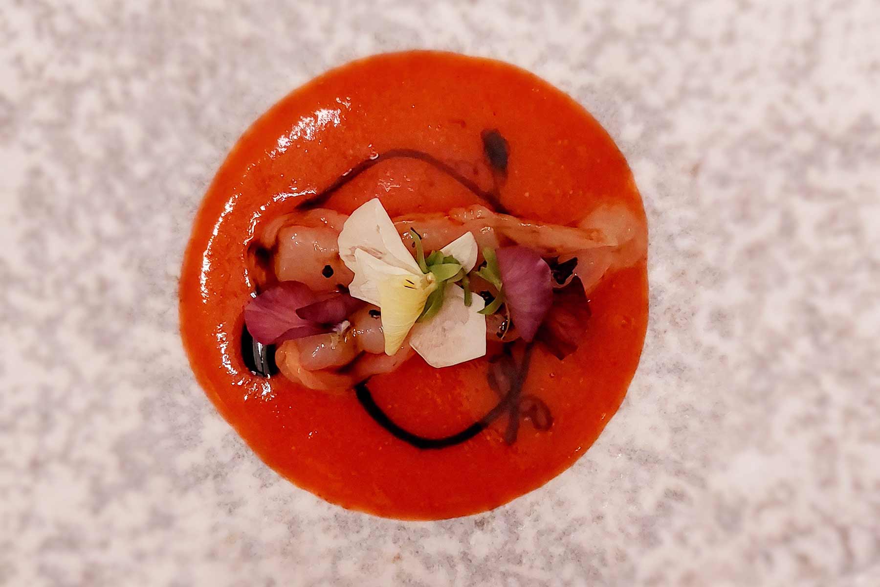 Runder Saucenspiegel aus Erdbeere mit zwei rohen Gambas, garniert mit essbaren Blüten.