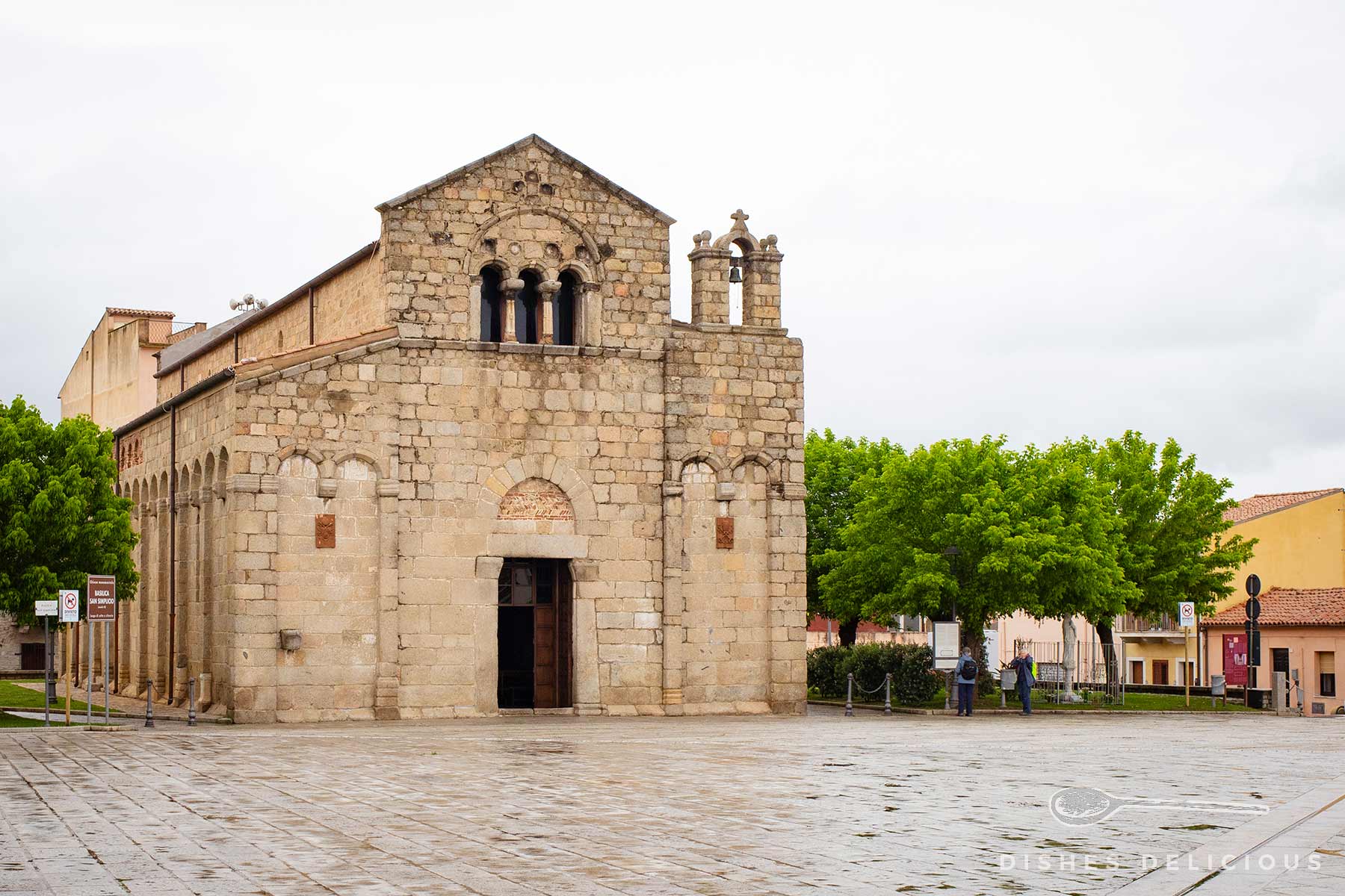 Romanische Kirche San Simplicio in Olbia mit steinerner Fassade, Rundbogenfenstern und kleinem Glockenturm auf einem gepflasterten Platz.