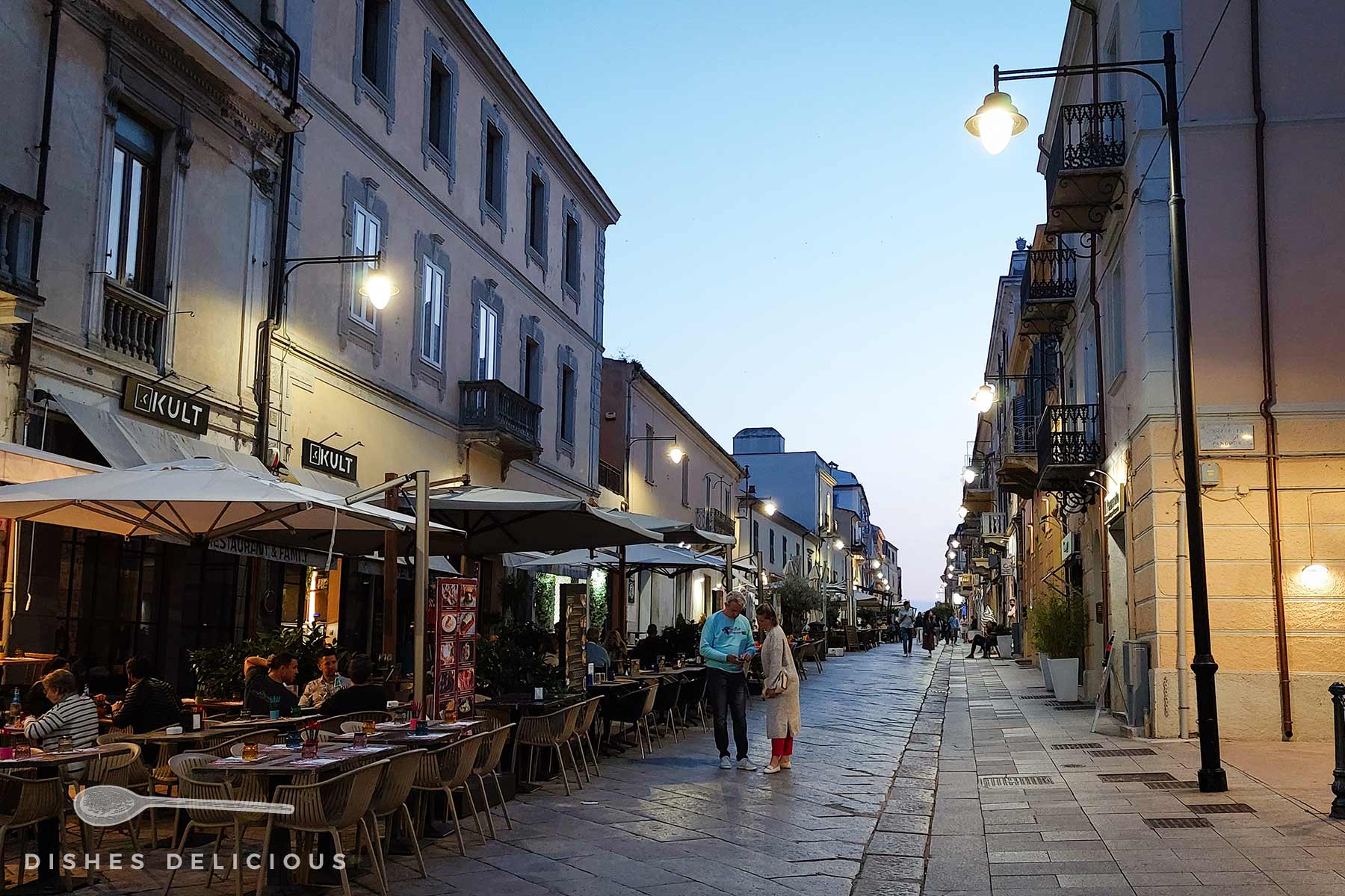 Abendliche Straßenszene in Olbia mit beleuchteten Cafés und Menschen, die auf dem gepflasterten Corso Umberto spazieren.