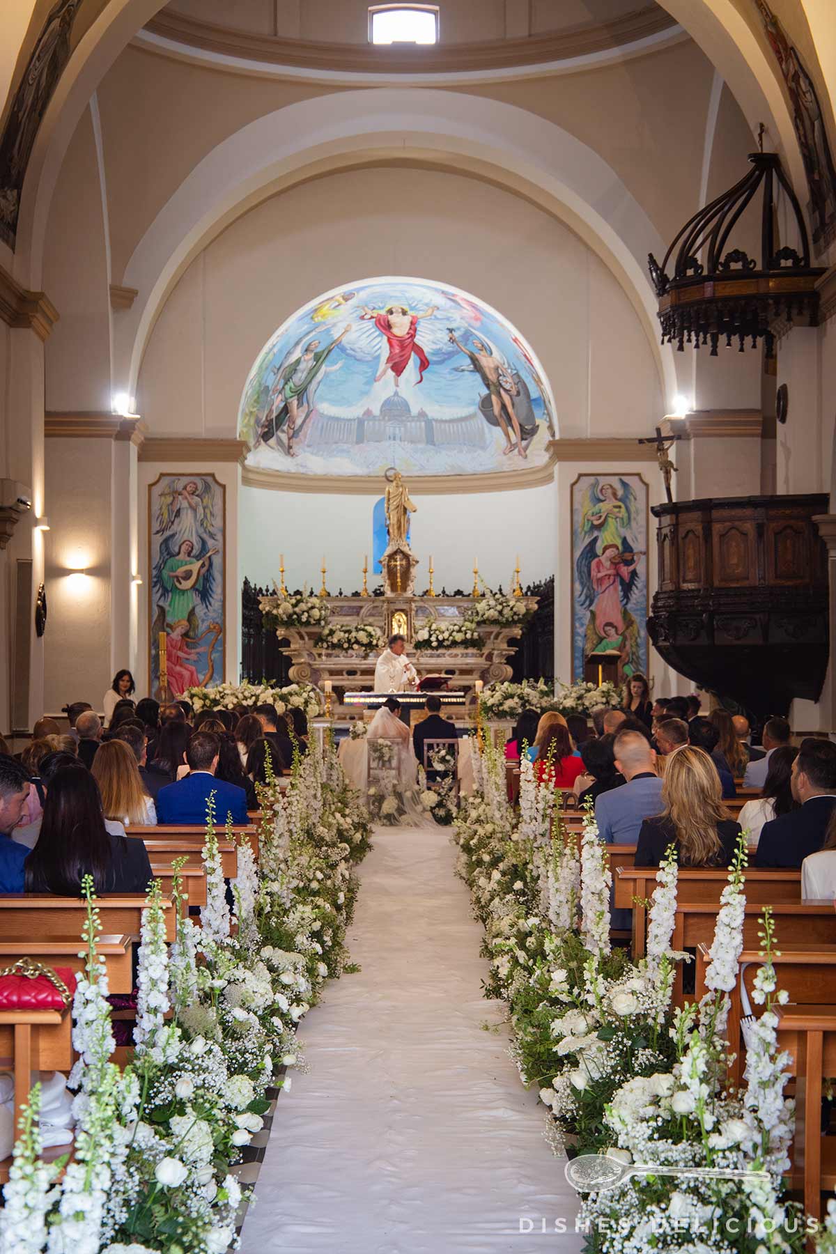 Blick auf den mit weißen Blumen geschmückten Mittelgang einer Kirche mit Altar, an dem ein Priester steht, und Gästen auf Holzbänken bei einer Hochzeit in der Chiesa San Paolo.