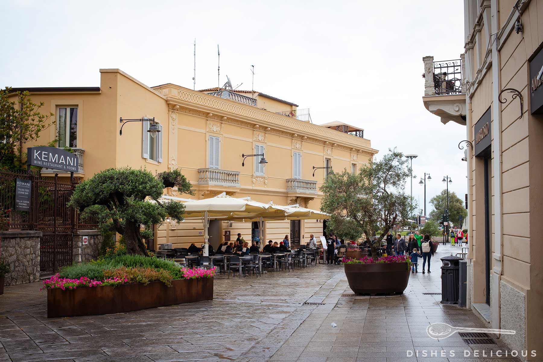 Corso von Olbia mit Außenbestuhlung vor gelbem Novecento-Palazzo und großen Pflanzkübeln mit Bäumchen und Blumen.