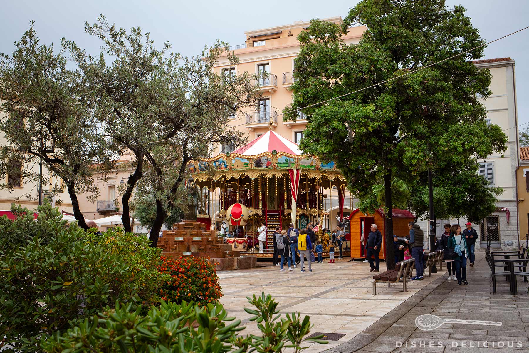 Karussell auf der Piazza Matteotti in Olbia mit Bäumen, Büschen und mehreren Personen, umgeben von mehrstöckigen Gebäuden.