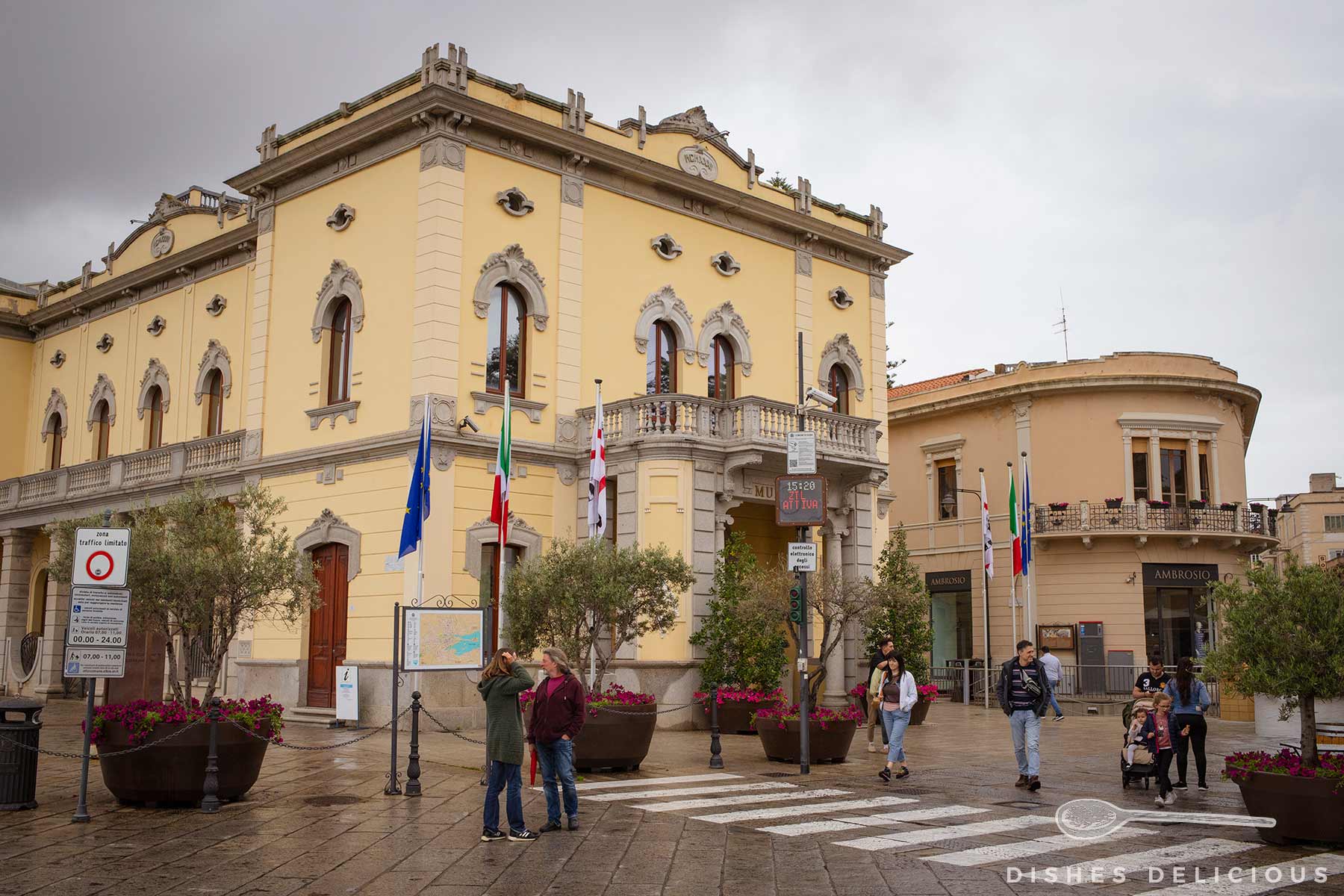 Historisches Rathausgebäude in Olbia mit gelber Fassade, mehreren Flaggen und Menschen auf dem gepflasterten Platz davor.