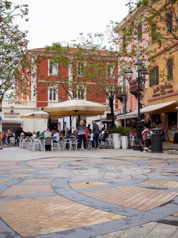 Piazza Margherita mit gemustertem Boden, Bäumen, Sitzbänken und Cafés mit Außenbestuhlung in Olbia.