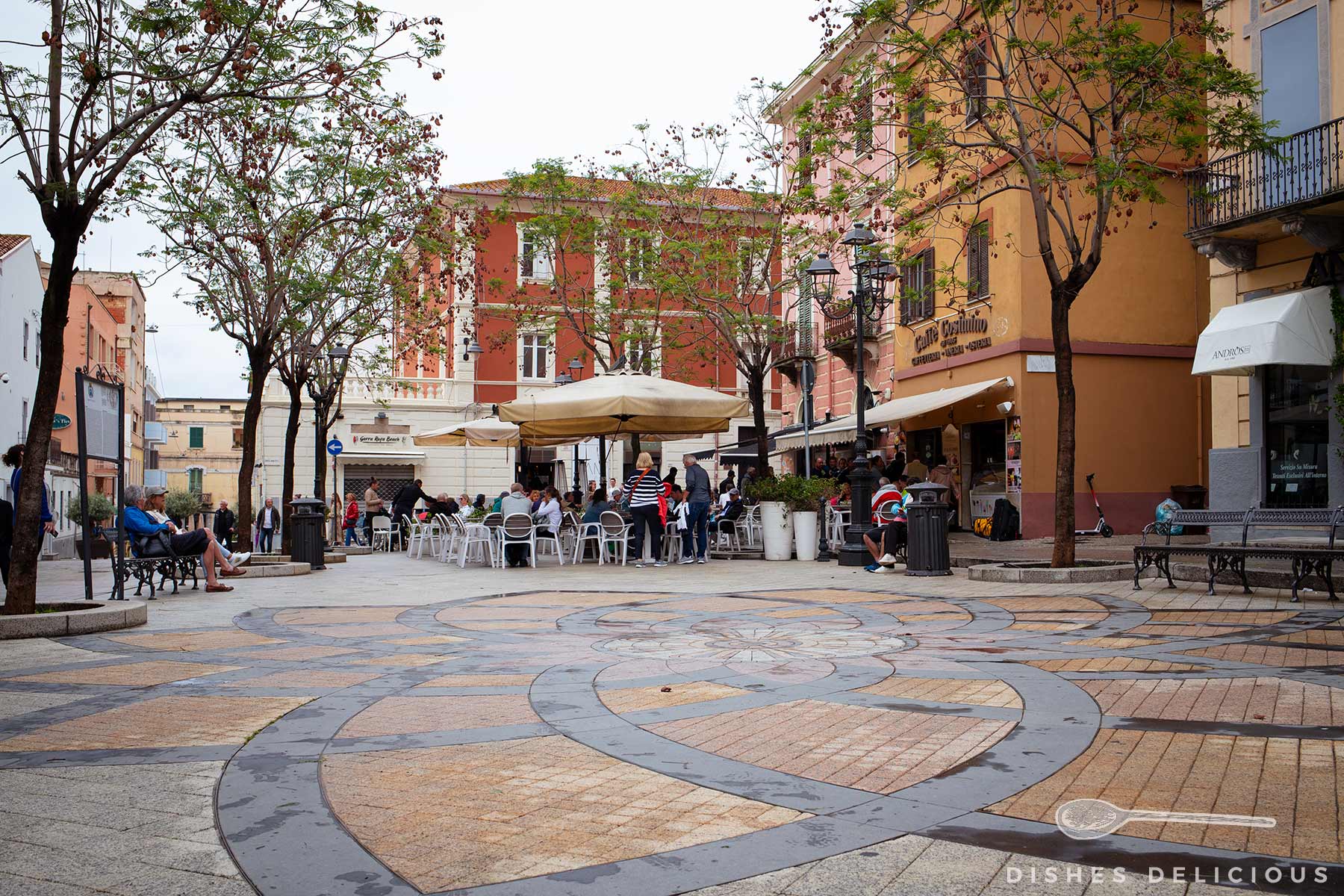 Piazza Margherita mit gemustertem Boden, Bäumen, Sitzbänken und Cafés mit Außenbestuhlung in Olbia.