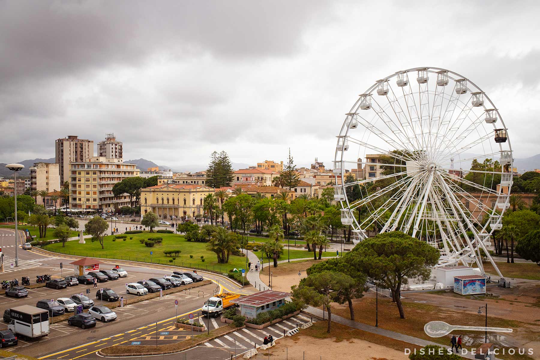 Großes Riesenrad am Rande der Altstadt von Olbia mit mehrstöckigen Gebäuden unter bewölktem Himmel im Hintergrund.