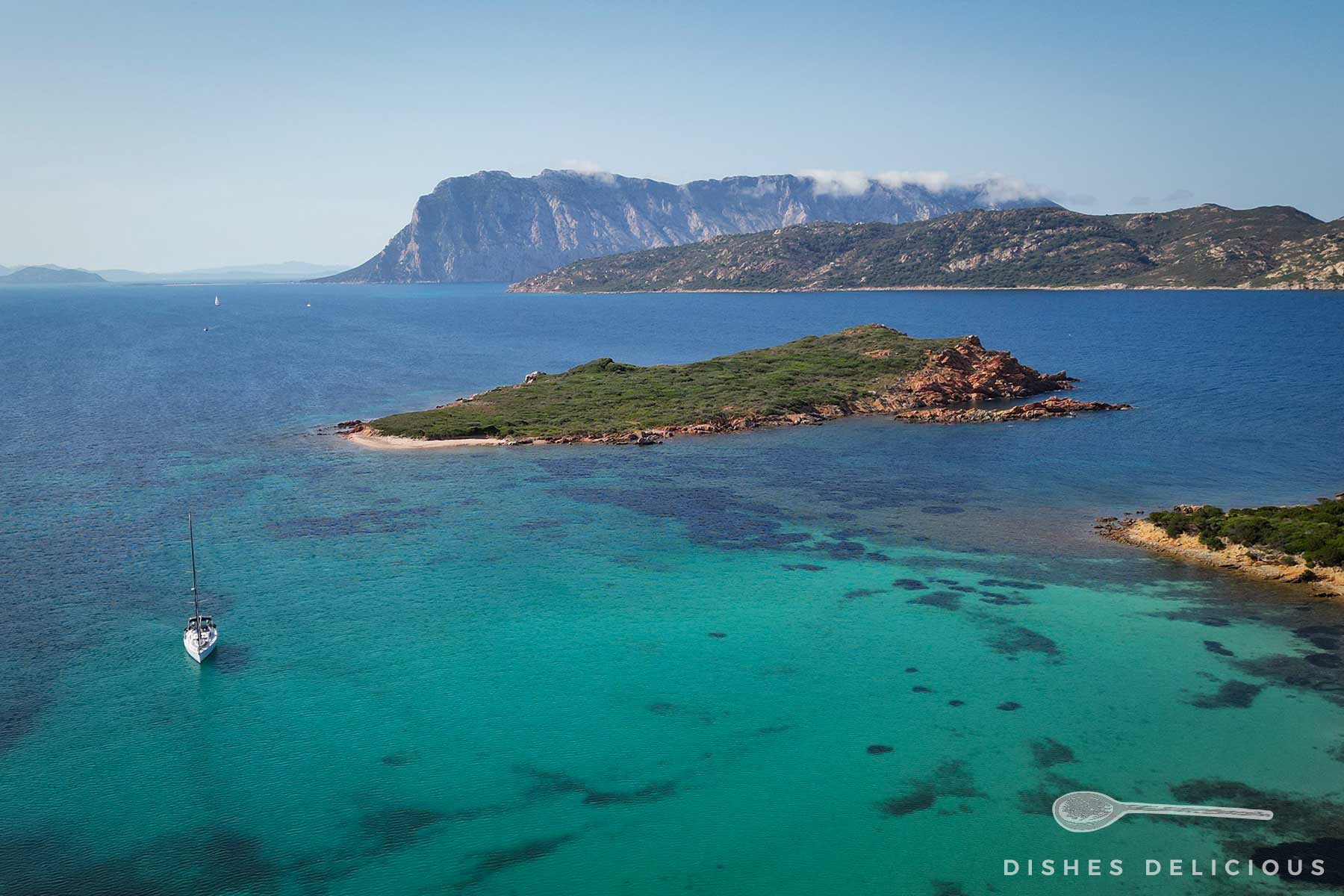 Blick auf die Inseln vor dem Capo Cavallo mit klarem türkisfarbenem Wasser, einem Segelboot und dem Tavolara-Berg im Hintergrund.